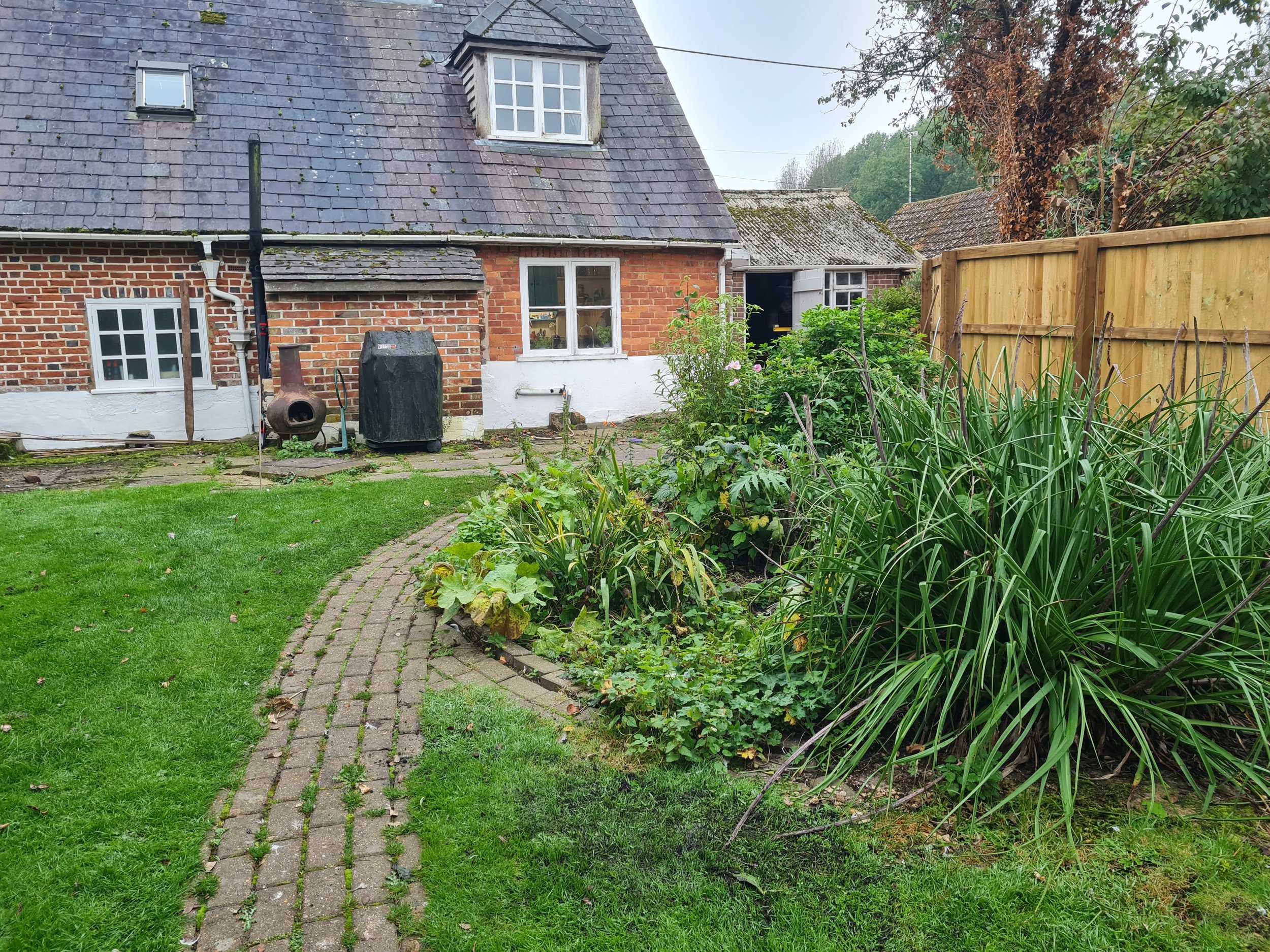 A backyard with a grassy lawn, a curved brick pathway, flower beds, and a wooden fence. In the background, there is a house with a brick and white painted wall, a sloped roof with moss and moss-growing chimney, and several windows. There are also garden plants and shrubs.
