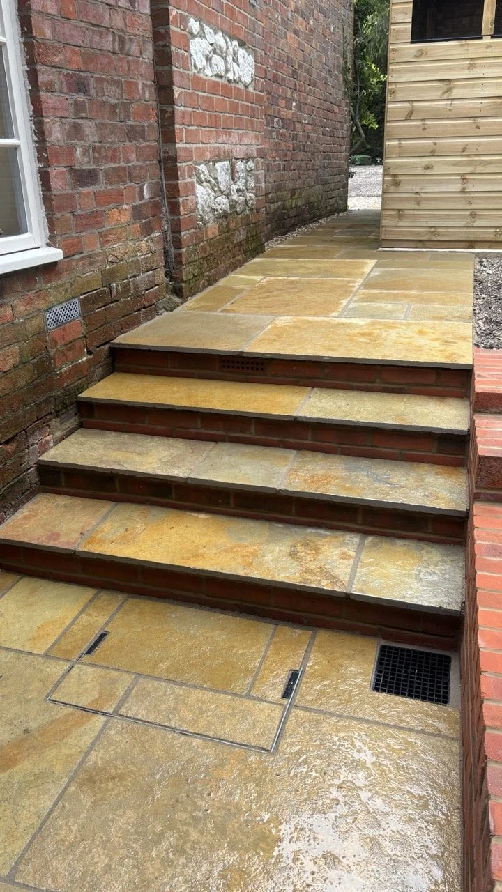 Stone step stairs outside a brick building, leading to a raised patio area, with drainage grates on the ground.
