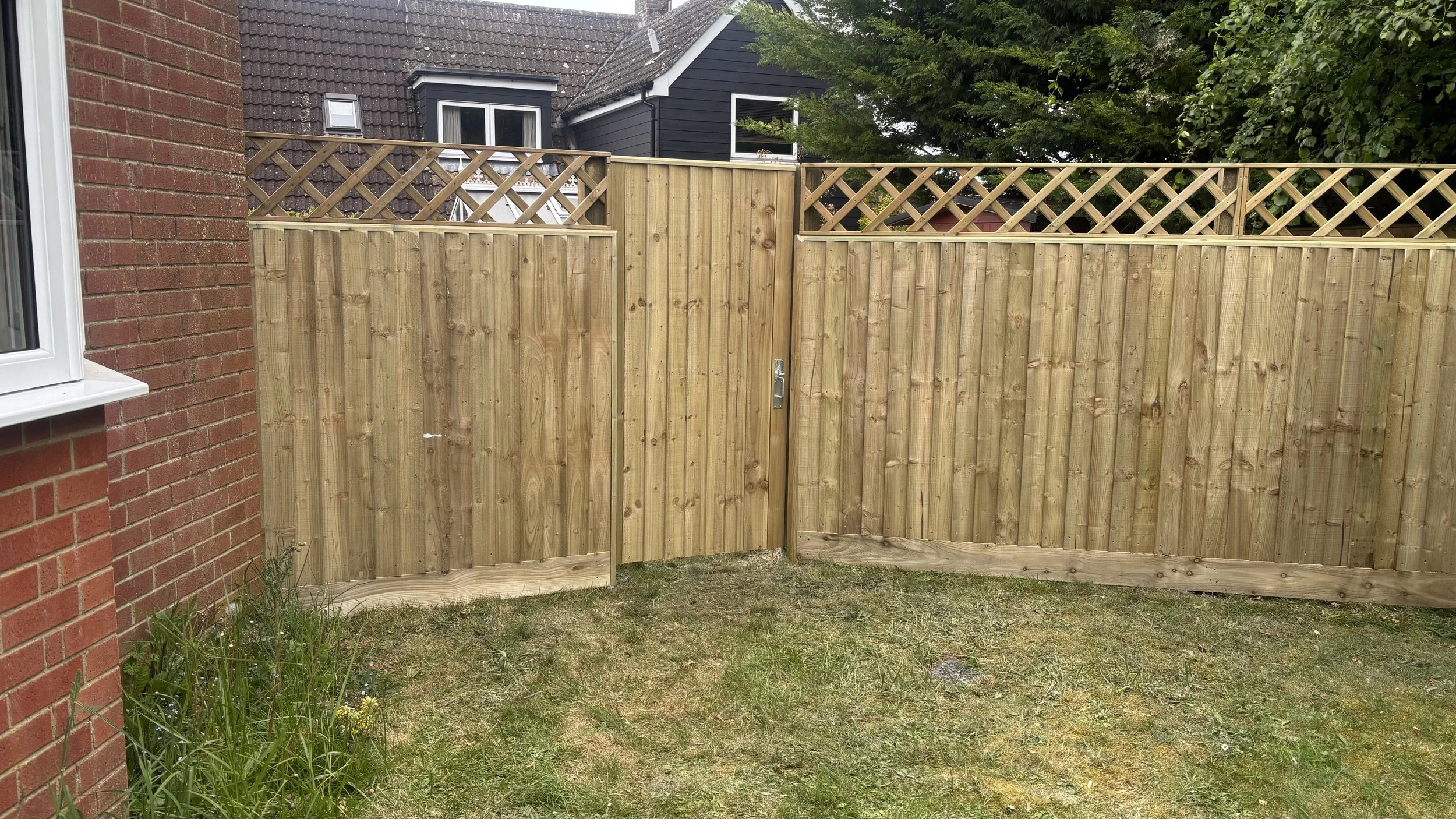 A wooden privacy fence with lattice top panels installed in a backyard, adjacent to a red brick house with a white-framed window, green grass, and some greenery and trees in the background.