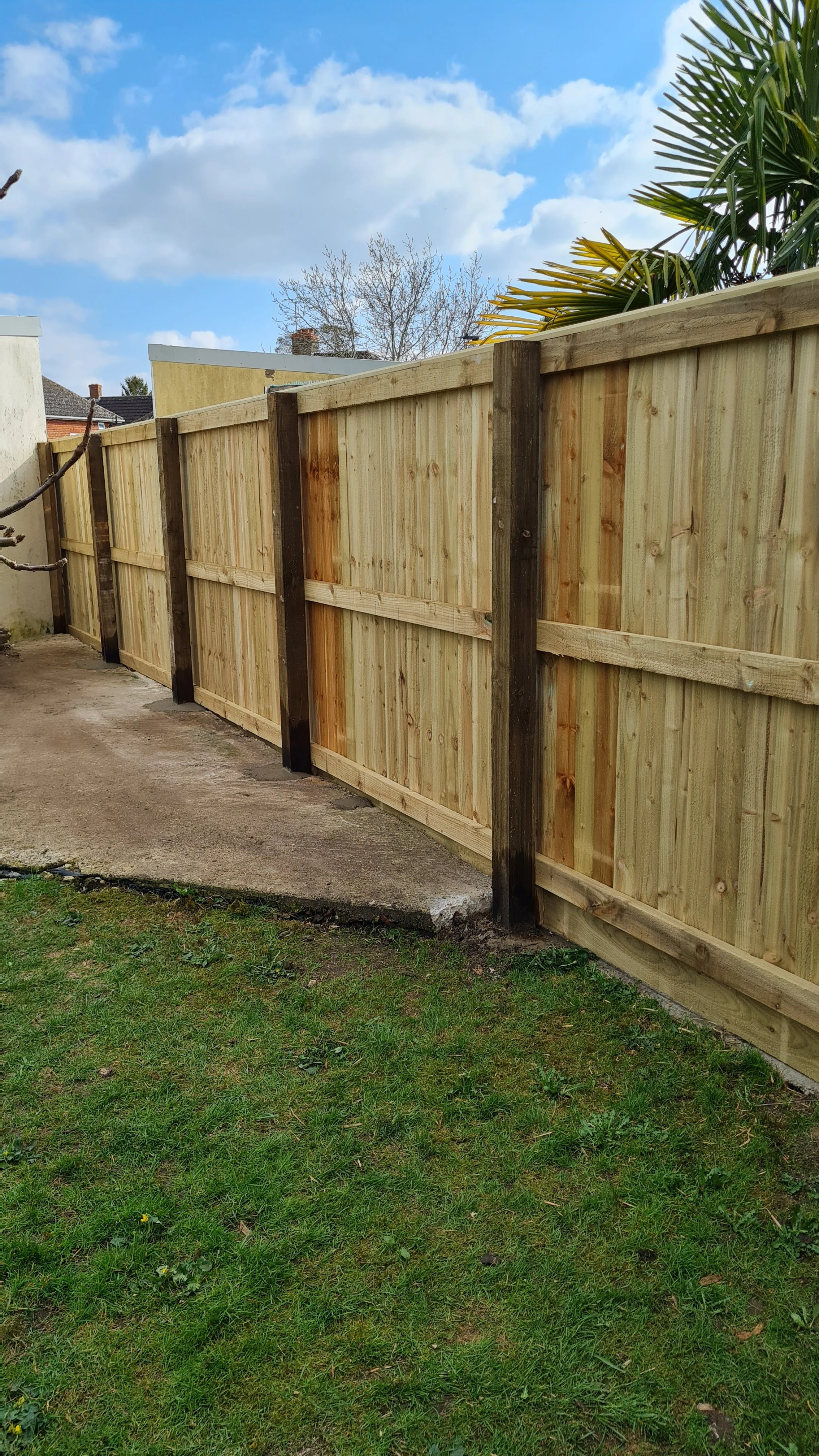 A newly built wooden privacy fence with vertical panels and dark support posts, installed along a concrete hard standing area and a lawn. The background includes a blue sky with scattered clouds and nearby houses.