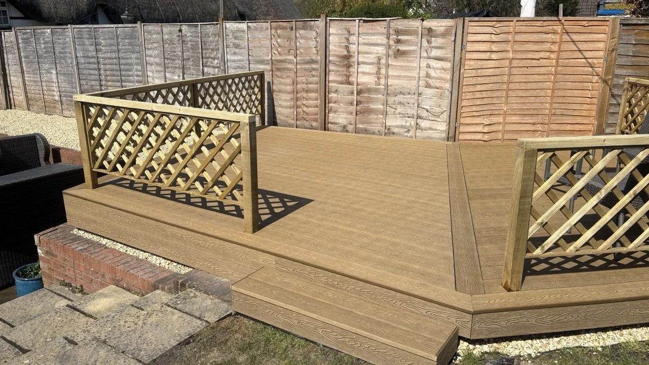 Newly built composite  deck with lattice Balustrade in backgarden, surrounded by a wooden fence. There are outdoor furniture pieces nearby and stone paving close to the deck.