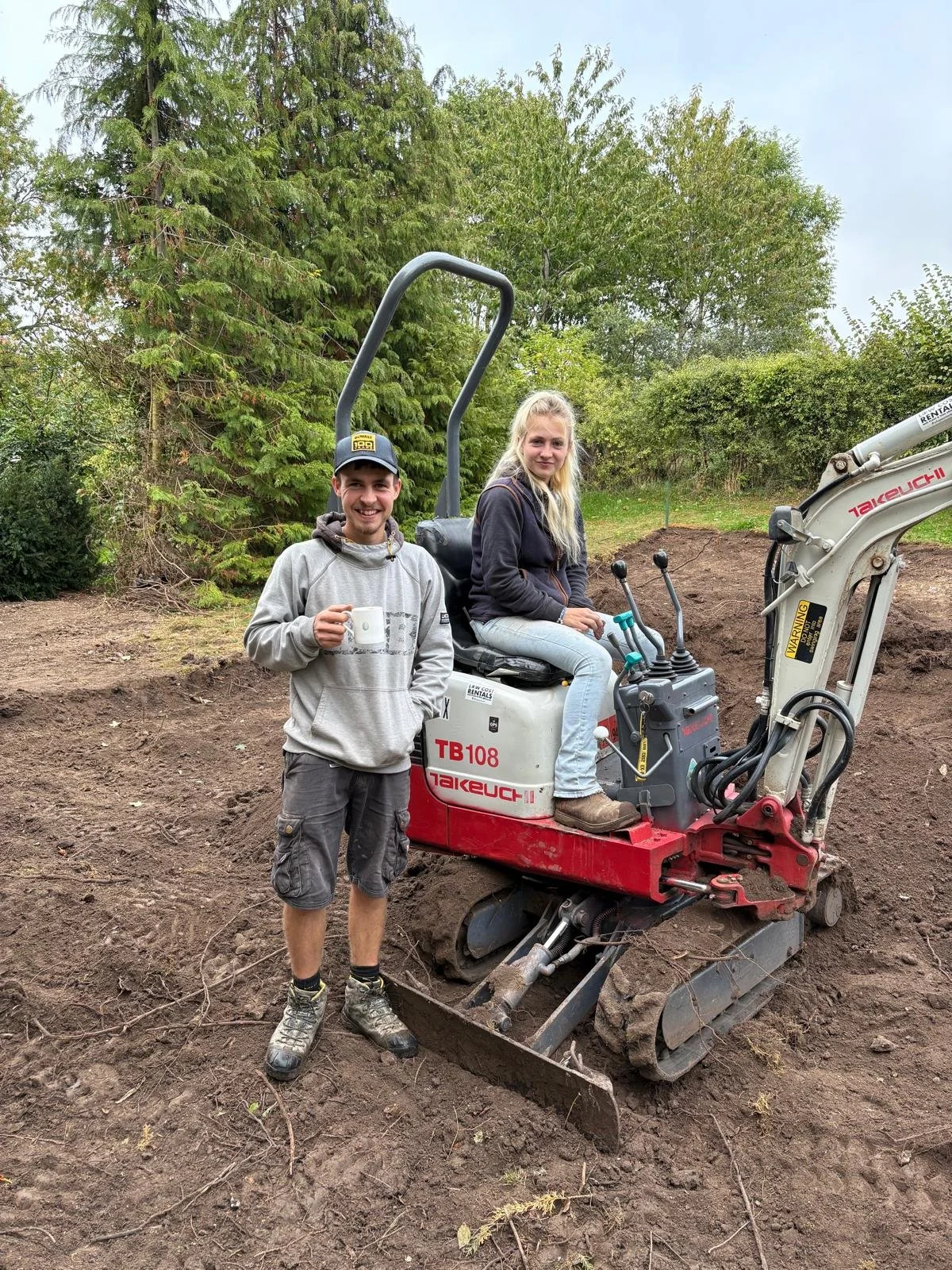 A man and a woman outdoors, with the woman sitting on a mini excavator and the man standing next to it, holding a coffee mug, surrounded by greenery and trees.