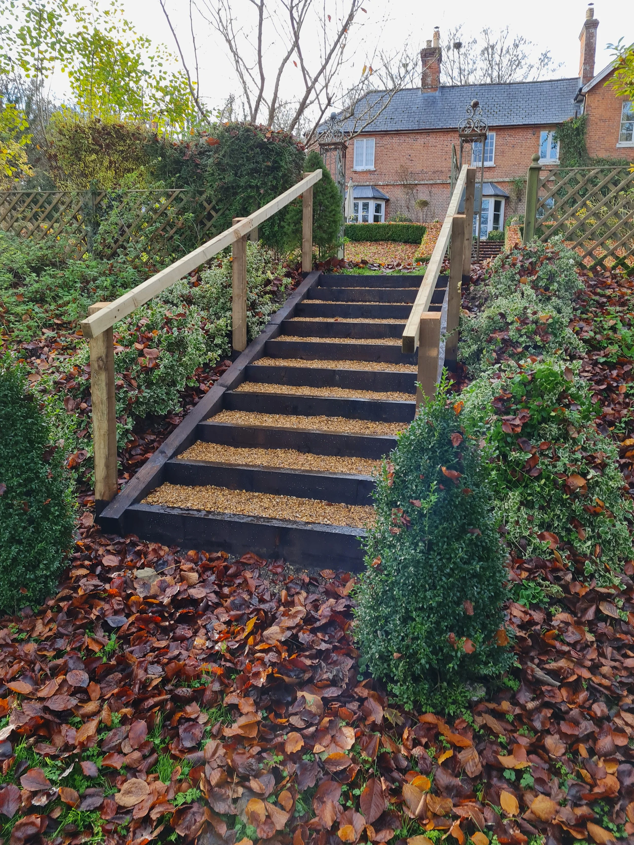 A garden staircase with black painted steps and wooden handrails, surrounded by fallen autumn leaves and lush green bushes, leading up to a brick house with chimneys and a garden in the background.