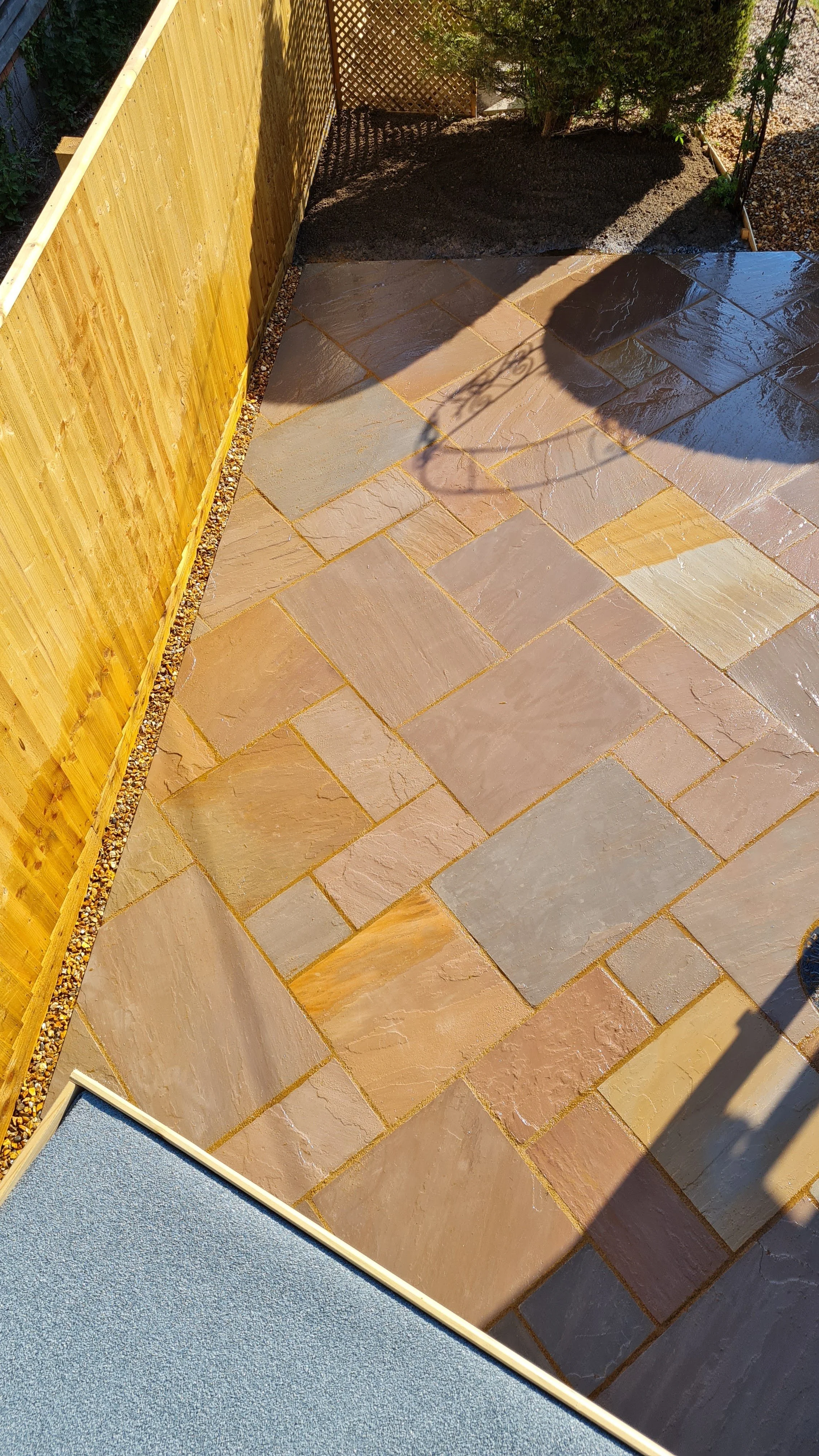 A backyard patio with multicolored stone tiles, a yellow wooden fence on the left, a small patch of dark soil with a bush on the upper right, and a shadow cast across part of the tiled floor.