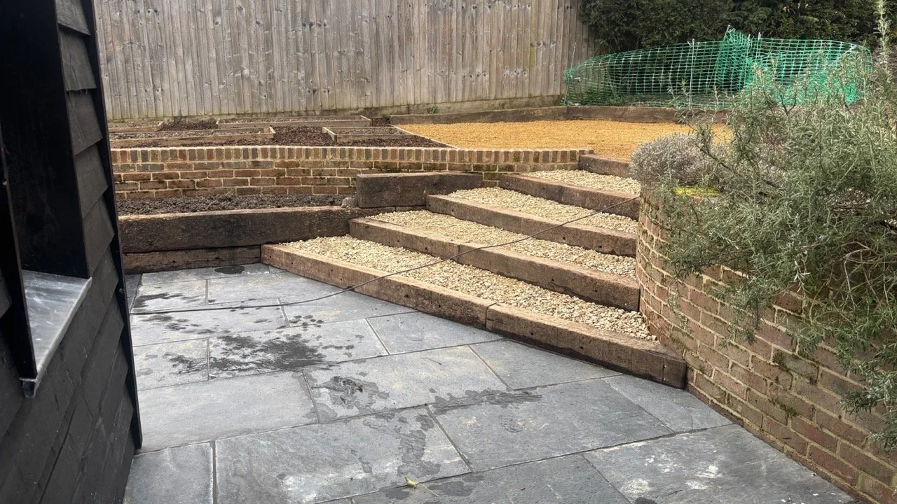 A backyard with stone paving and wooden steps leading up to a raised garden bed with gravel, soil, and a green netted area for planting, enclosed by a wooden fence.