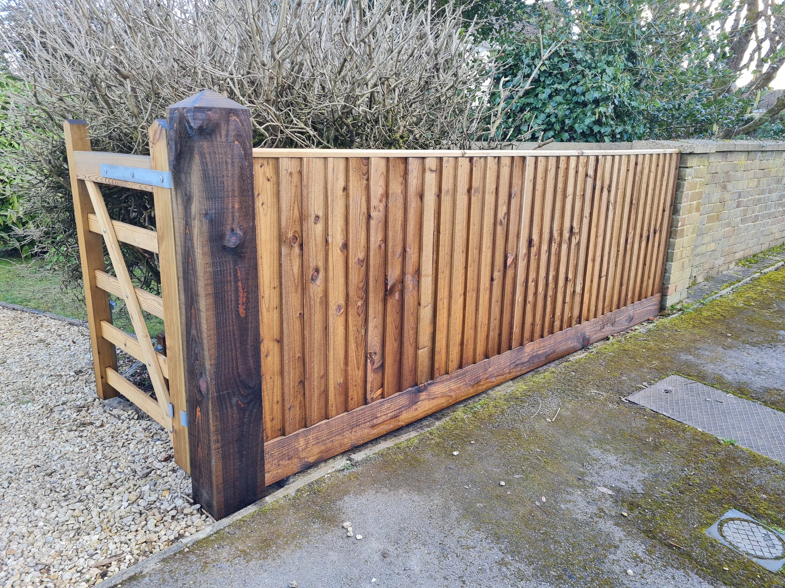 A newly installed wooden fence with upright slats runs along a garden boundary, with a brick wall on one side and leafless shrubs in the background. There is a gravel path and mossy ground in the foreground.