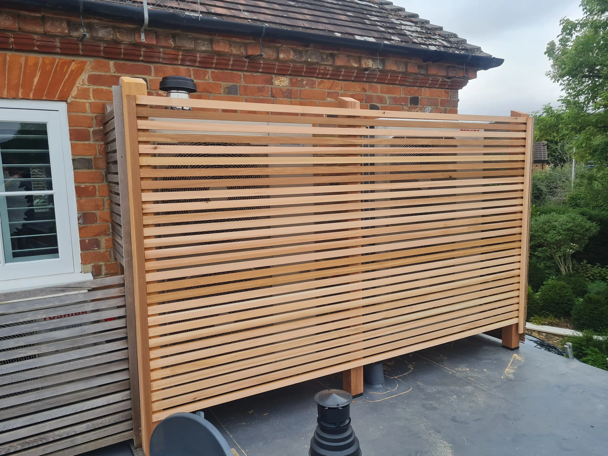 A Cedar screen installed around aircon units on a flat roof of a brick house with a small window. Part of a black drainage pipe and outdoor equipment are visible, with trees and cloudy sky in the background.