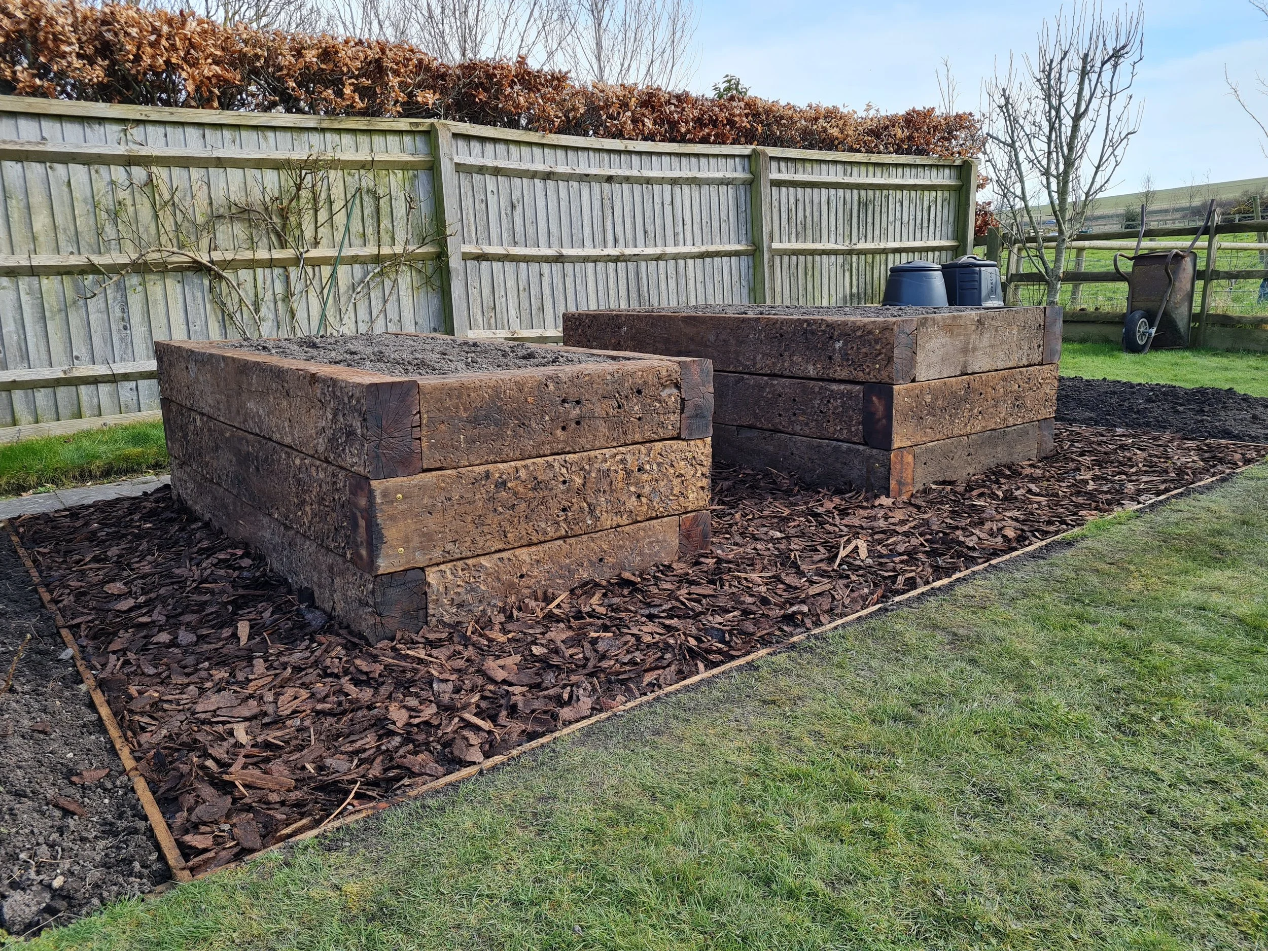 Two large wooden planters filled with soil, situated in a garden area with mulch on the ground. A wooden fence with some dried vines growing on it and a few trees in the background. Gardening tools and containers are also visible.