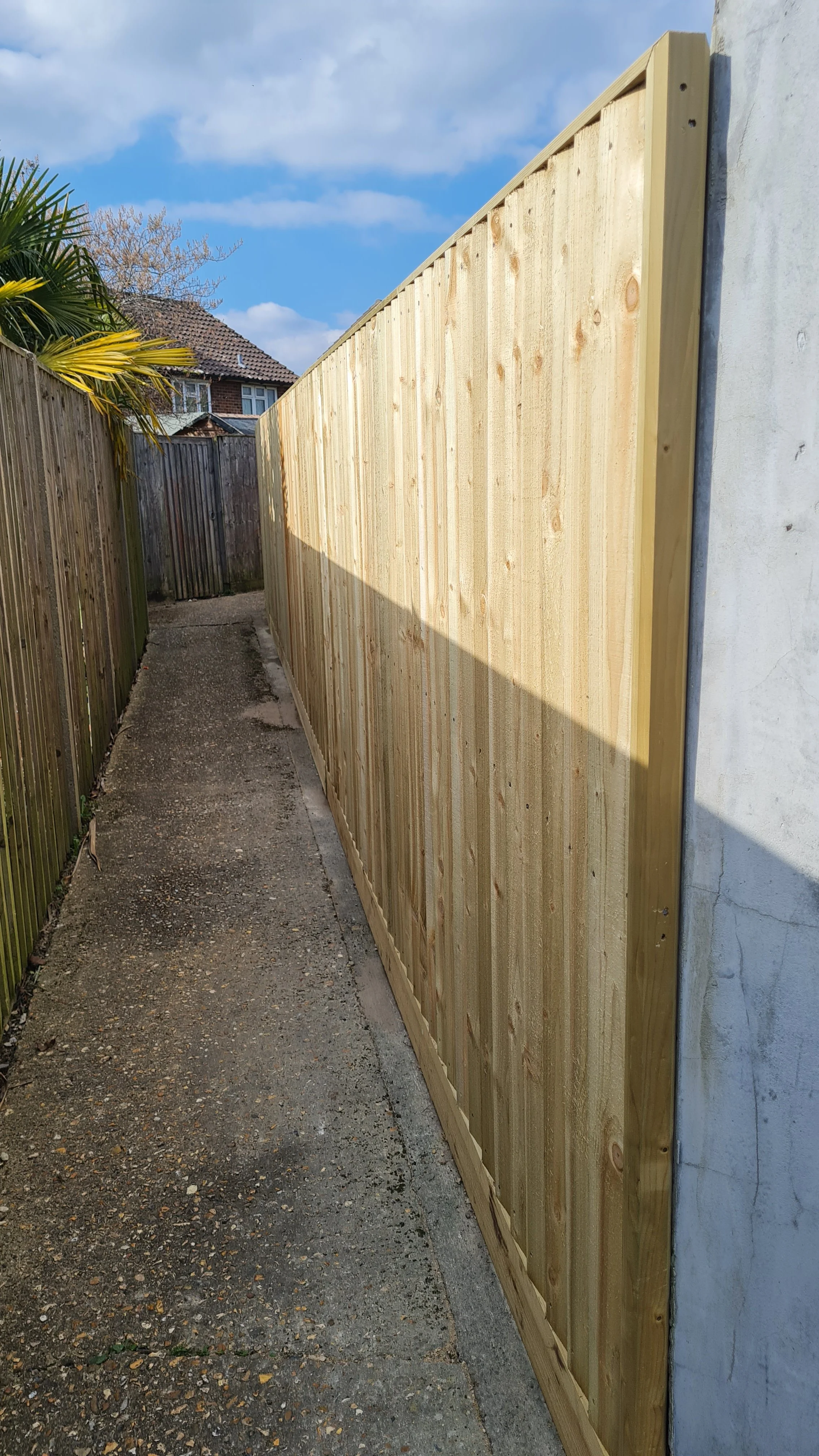 New wooden fence installed along a narrow concrete pathway between a house and a wooden fence, with a clear blue sky and nearby houses in the background.