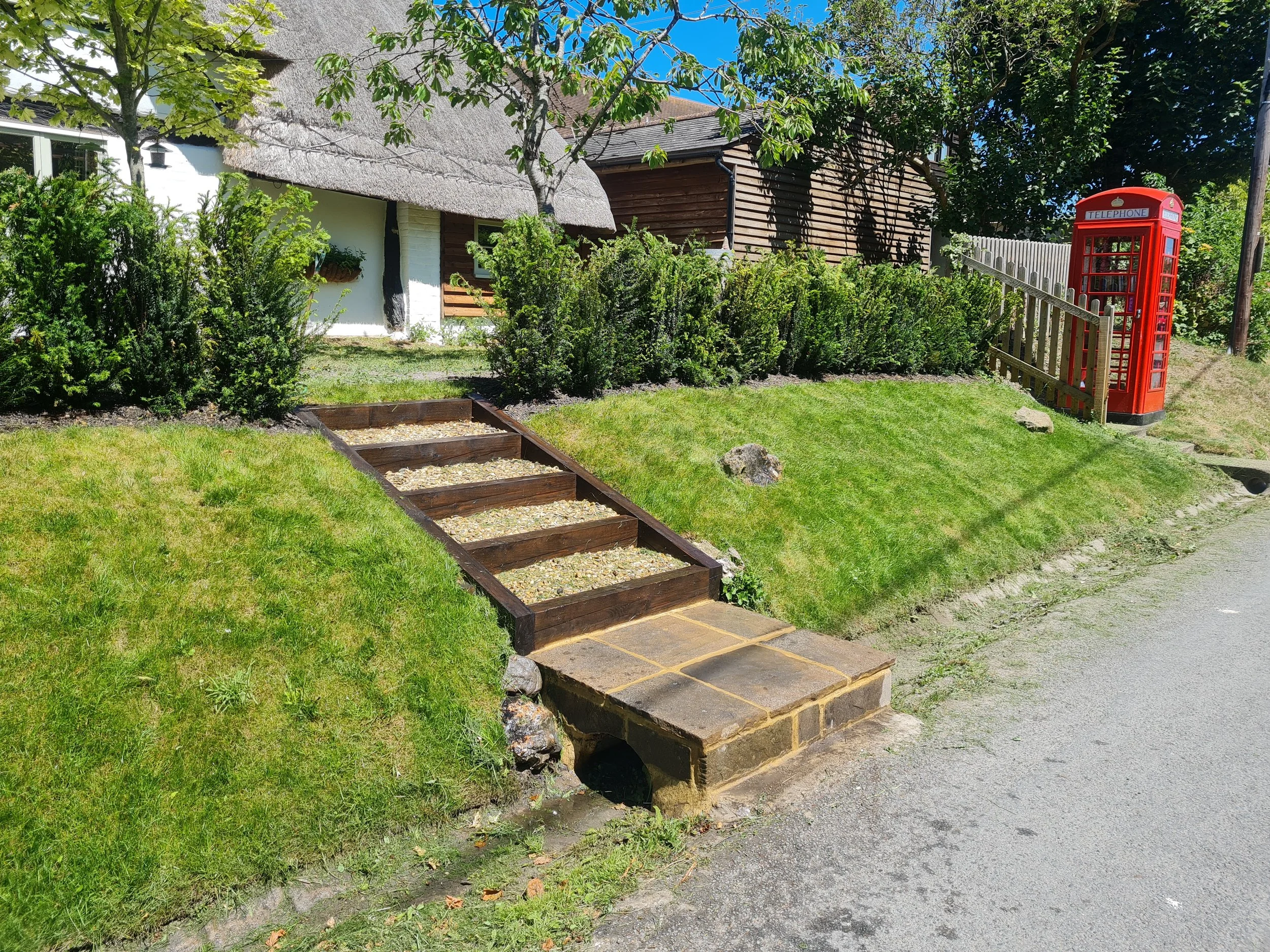 A garden with green grass and bushes, a stone pathway, a white house with a thatched roof, a wooden shed, a red telephone booth, and trees with green leaves under a clear blue sky.