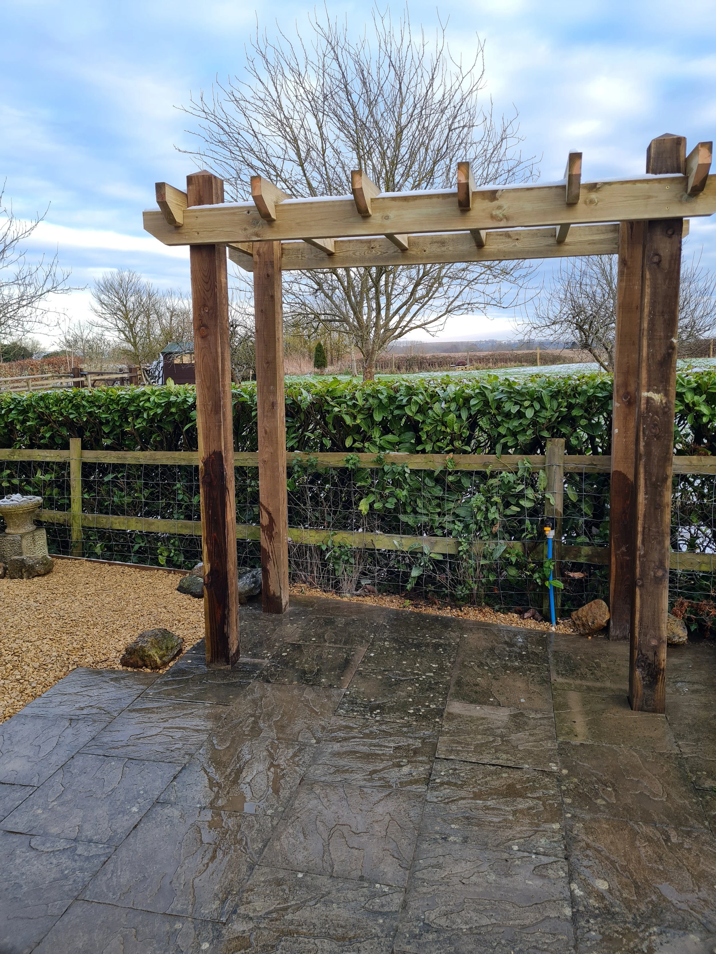 A backyard scene with a wooden garden arch structure, some trees, a hedge, and a distant field under a cloudy sky.