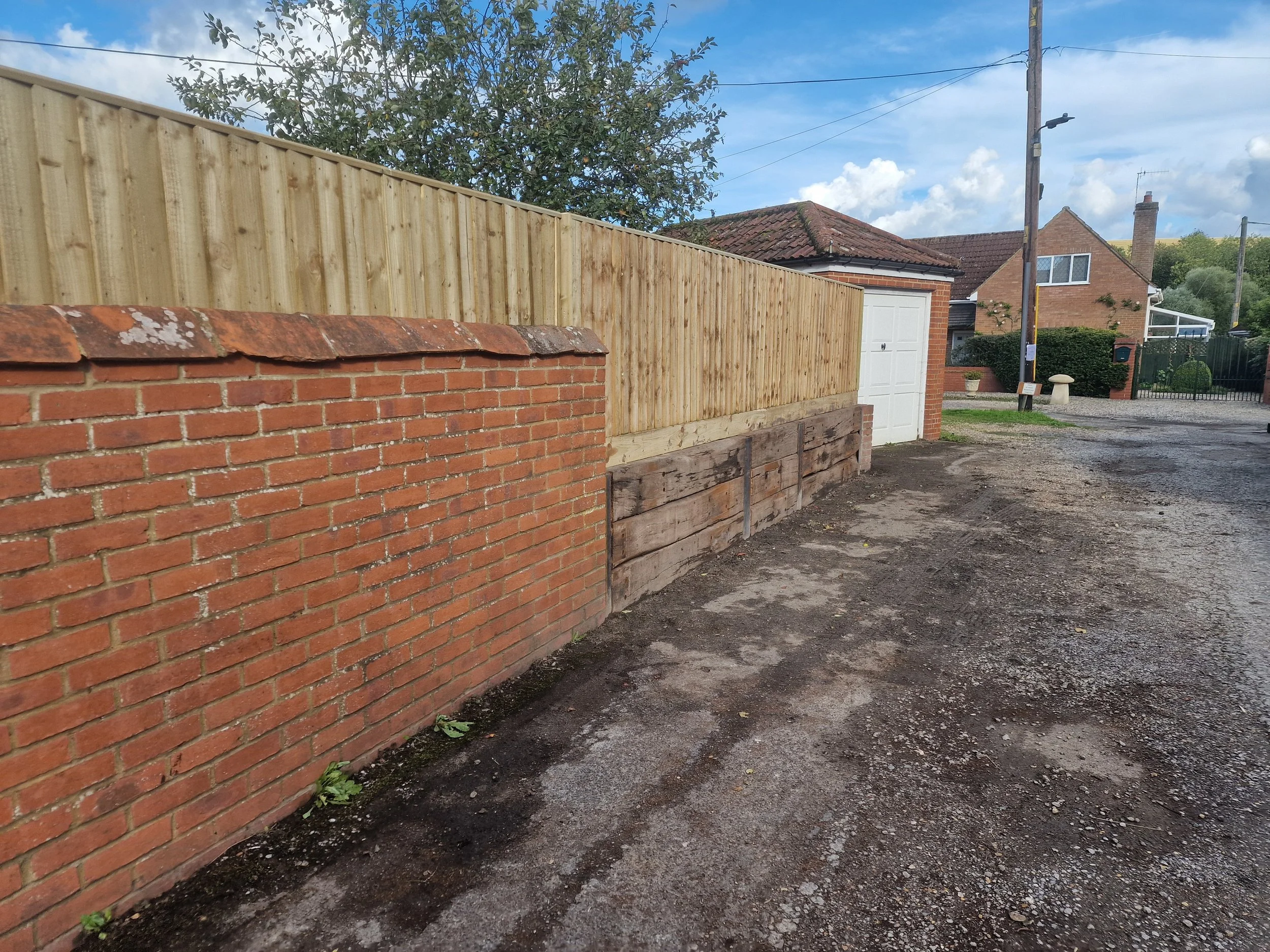 A newly constructed wooden fence runs along the side of a property, with some areas finished and other sections still under construction, next to a brick wall and driveway. In the background, there are houses, trees, power lines, and a partly cloudy 