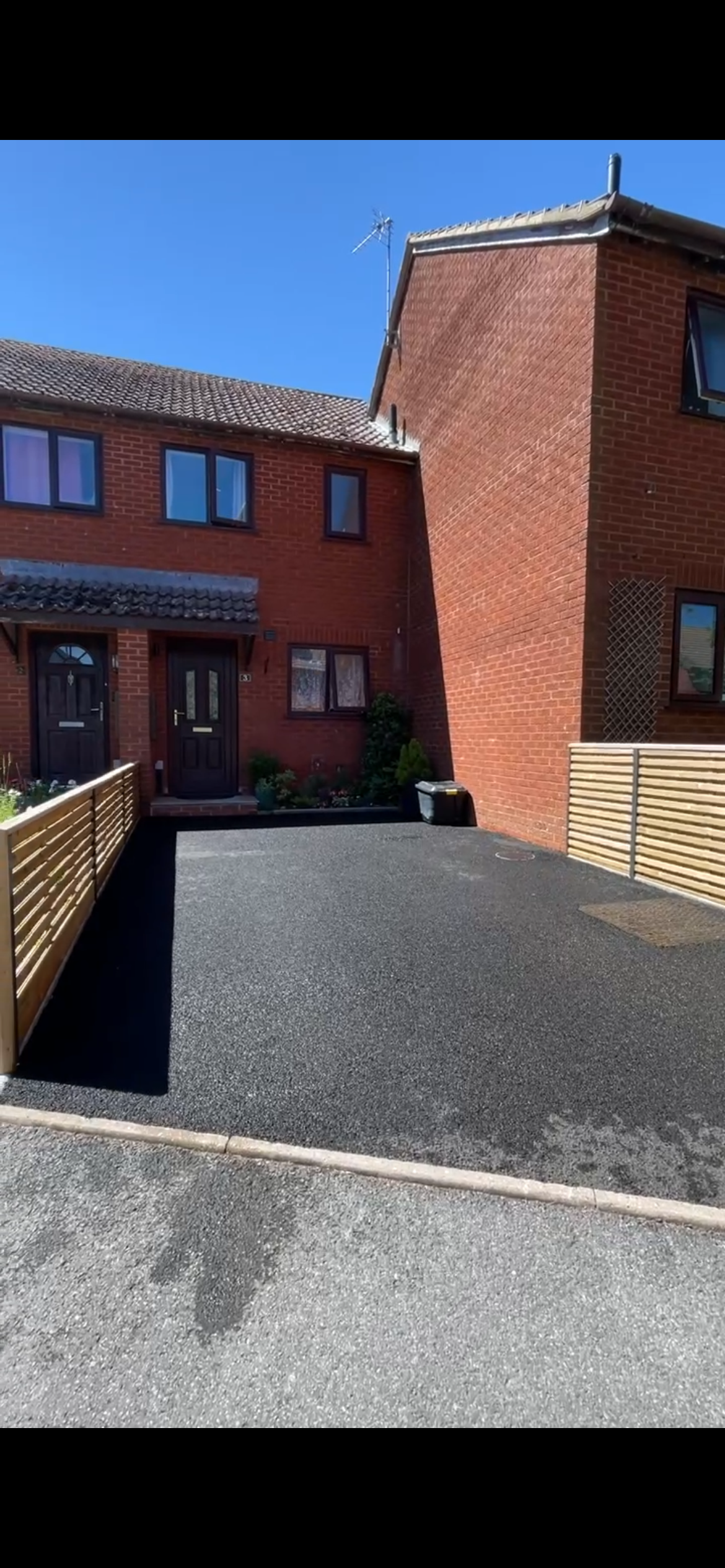 A clean driveway in front of two red brick townhouses, with two front doors, small garden beds, and wooden fences on each side, under a clear blue sky.