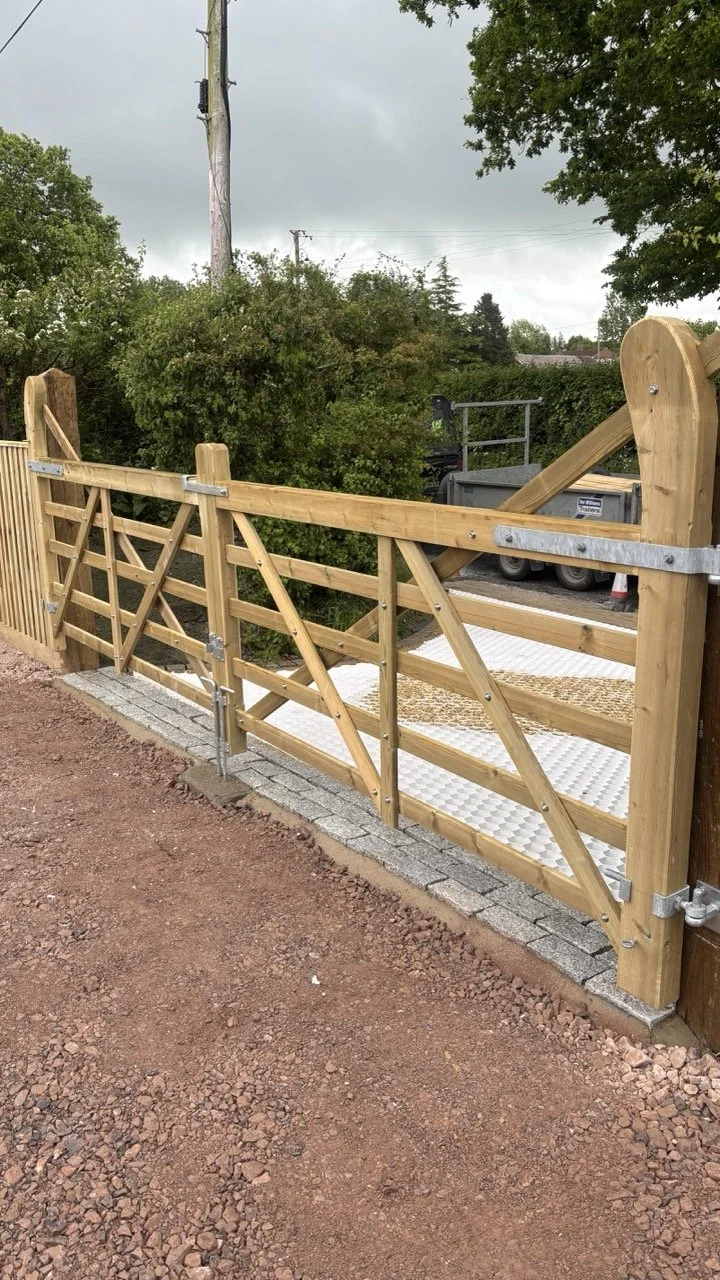Wooden gate with metal hinges and latch, part of a fence, leading to a gravel pathways and an area with a trailer or small vehicle, surrounded by trees and bushes.