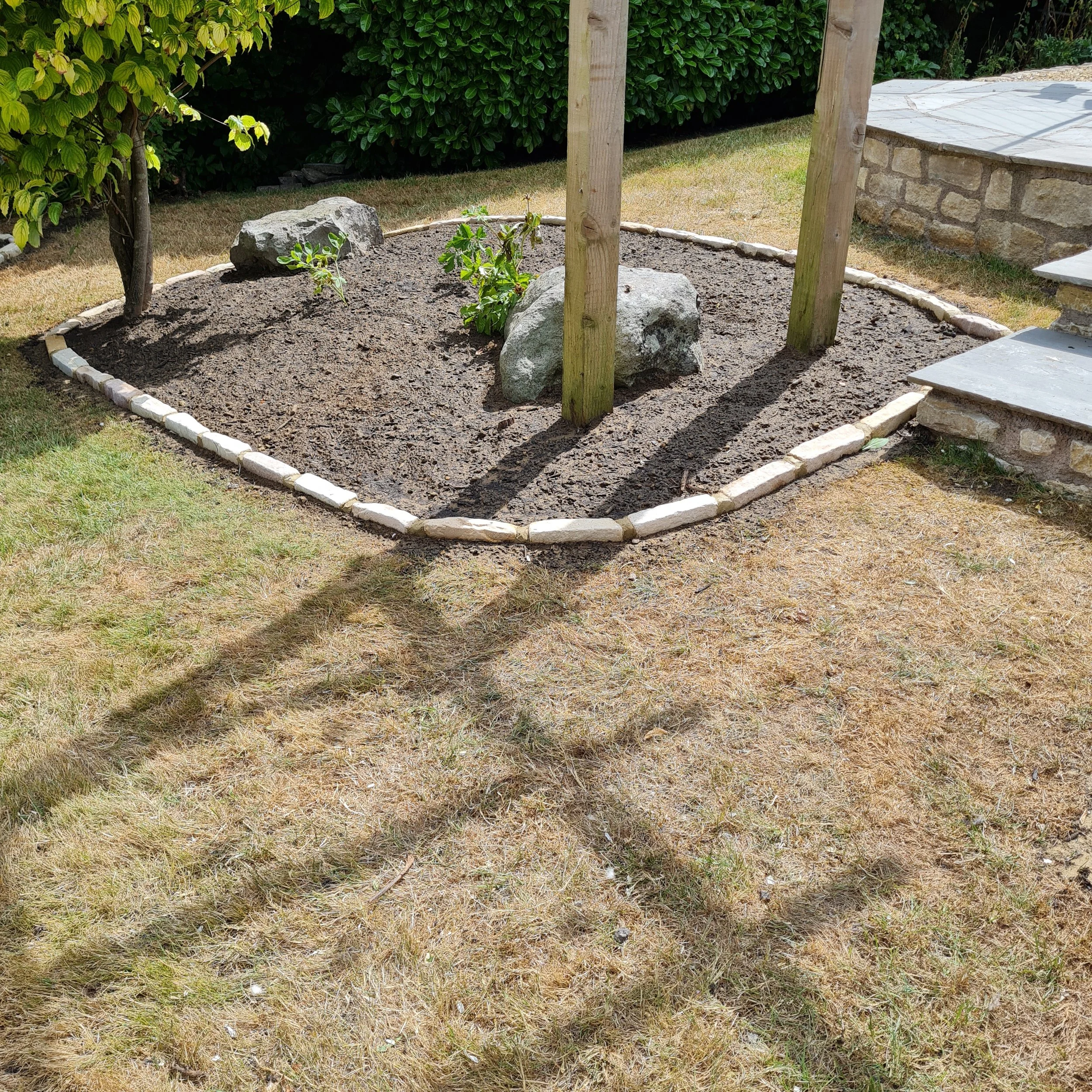 A small garden bed with dirt, a few young plants, and large rocks, surrounded by grass and some green bushes, with a stone patio and stairs on the right.
