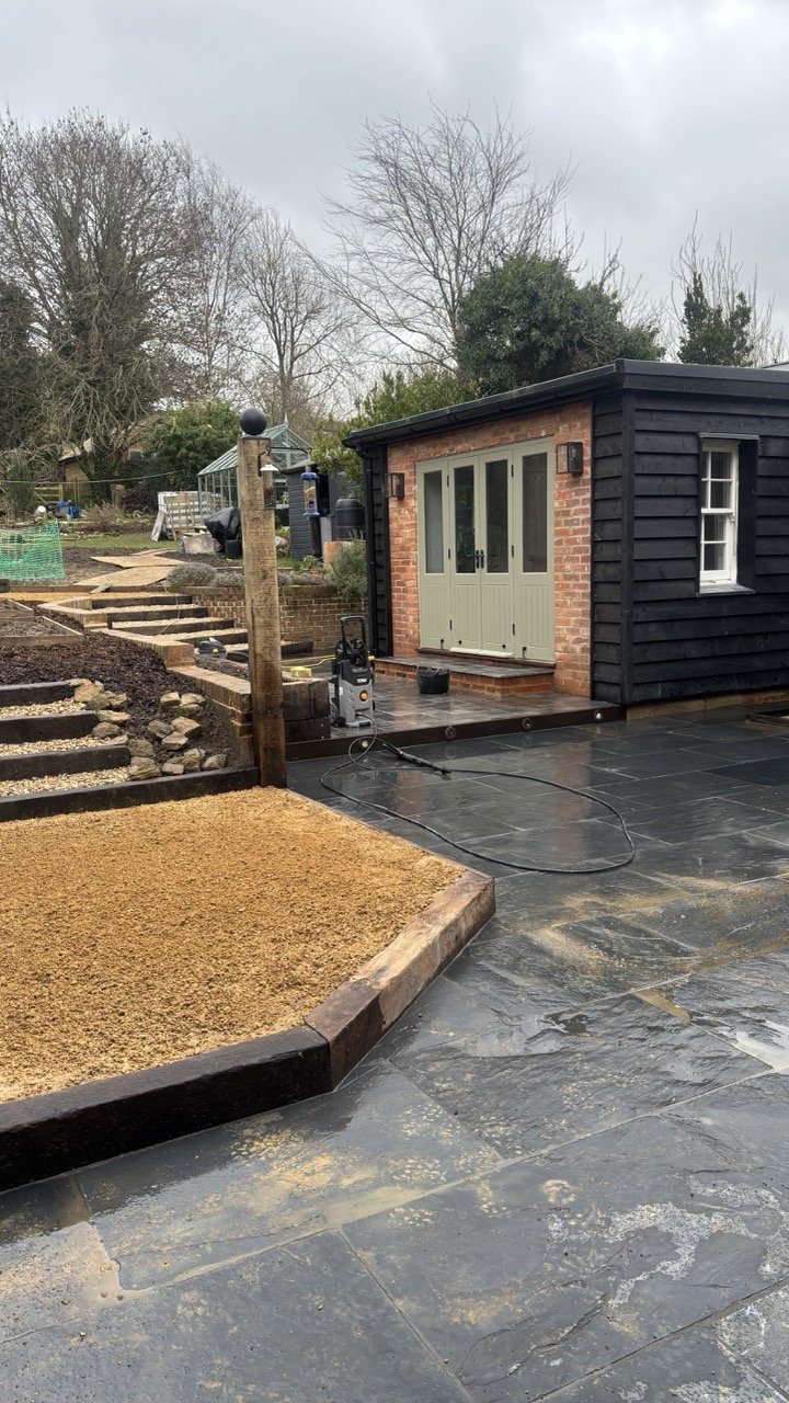 A backyard patio with black stone tiles and a small brick and black wooden shed with a double door and window, adjacent to a garden area with gravel, brick borders, and steps leading up a hill in a cloudy outdoor setting.