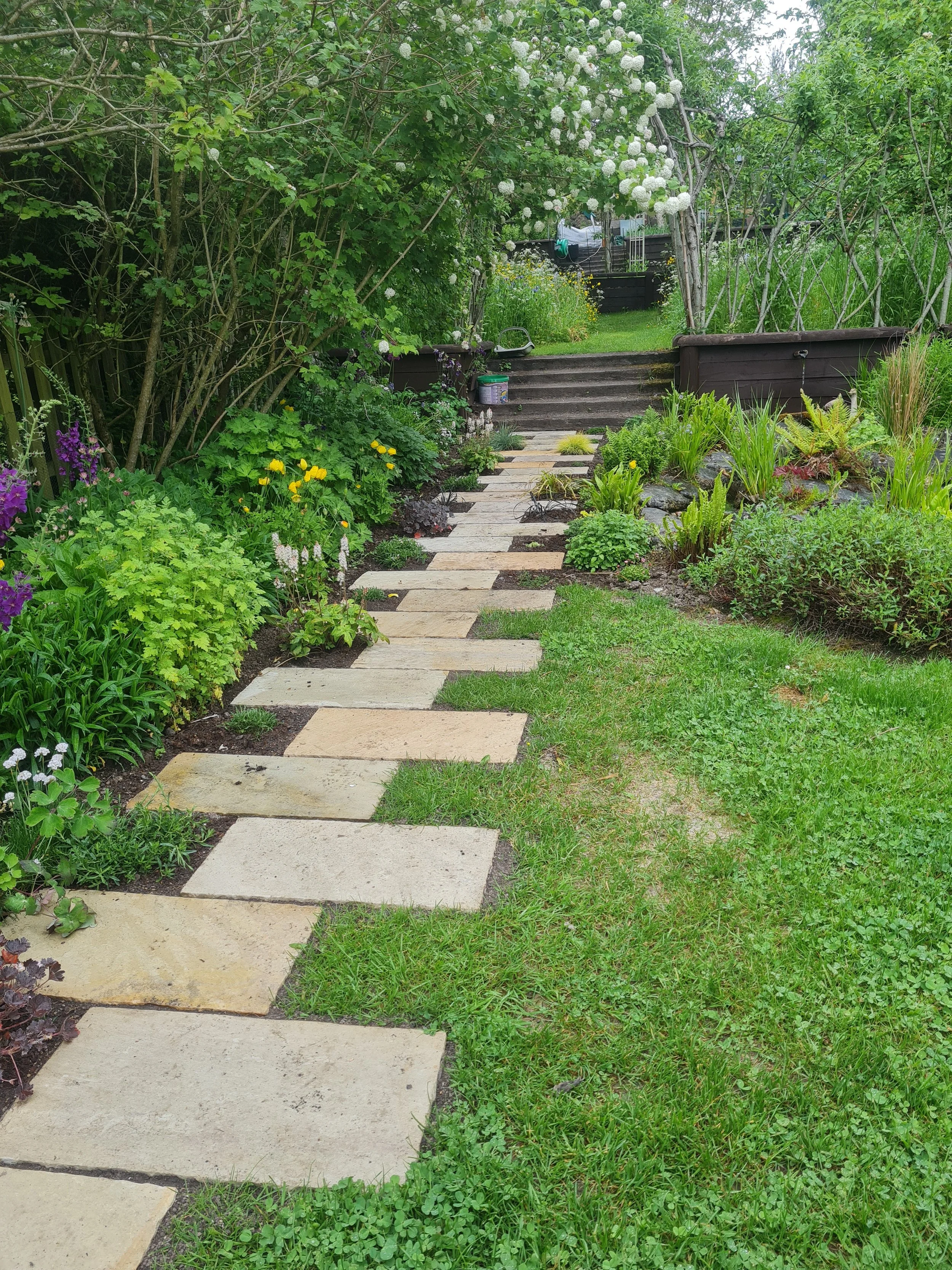 A garden pathway made of rectangular stone slabs leading up a small set of stairs through green plants and flowers, with a floral archway overhead.