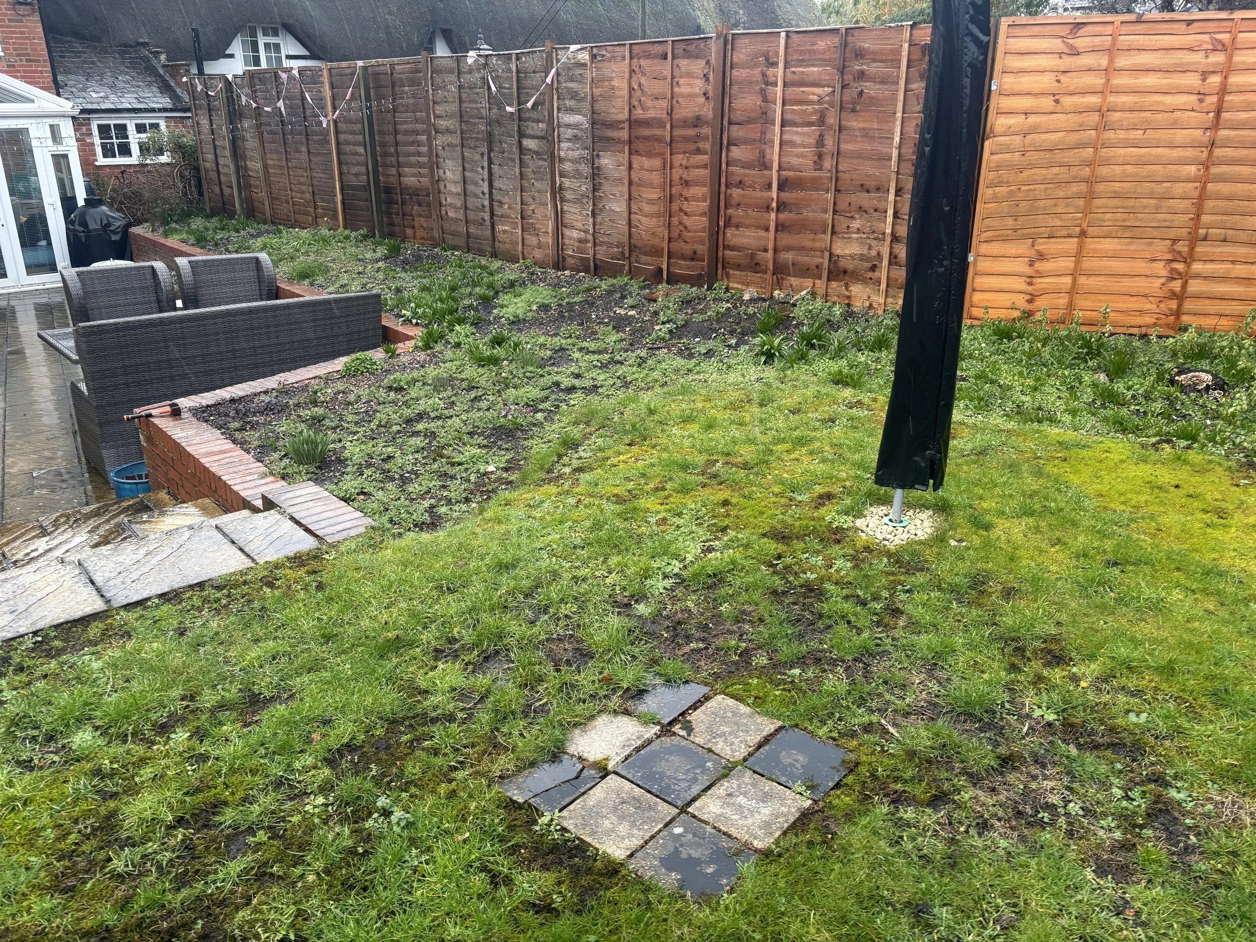 A backyard garden with a wooden fence, a small brick border, a garden bed, patio furniture, and some paving stones. The weather appears to be rainy and cloudy.