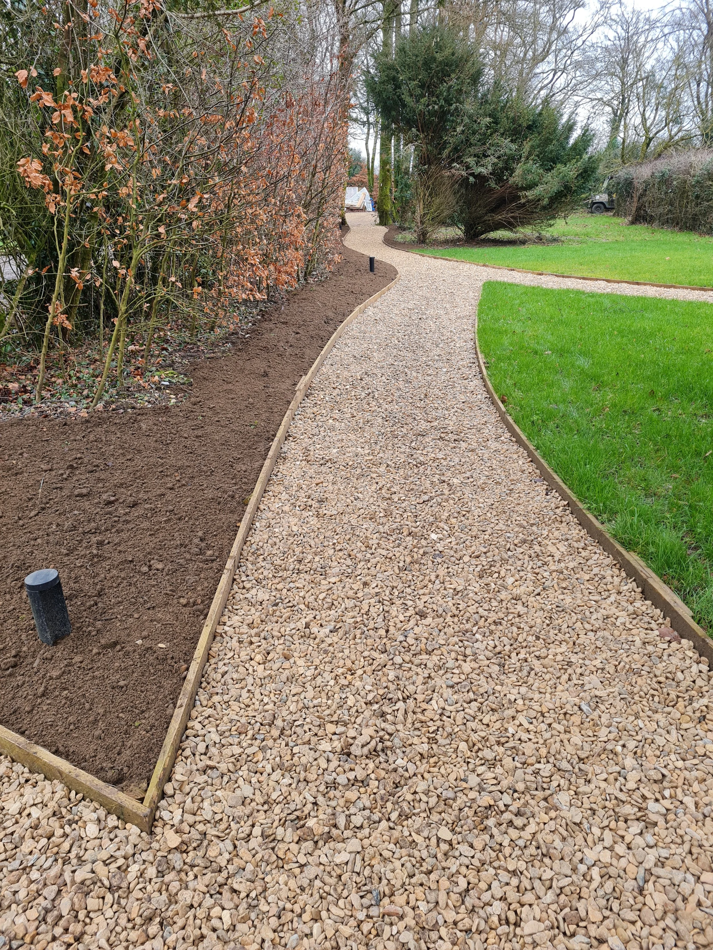 A gravel pathway curves through a yard, with new wooden edging on both sides. On the left, there is a flower bed with dark soil and some small plants. On the right, there is lush green grass. Tall bushes and trees line the background, with some trees