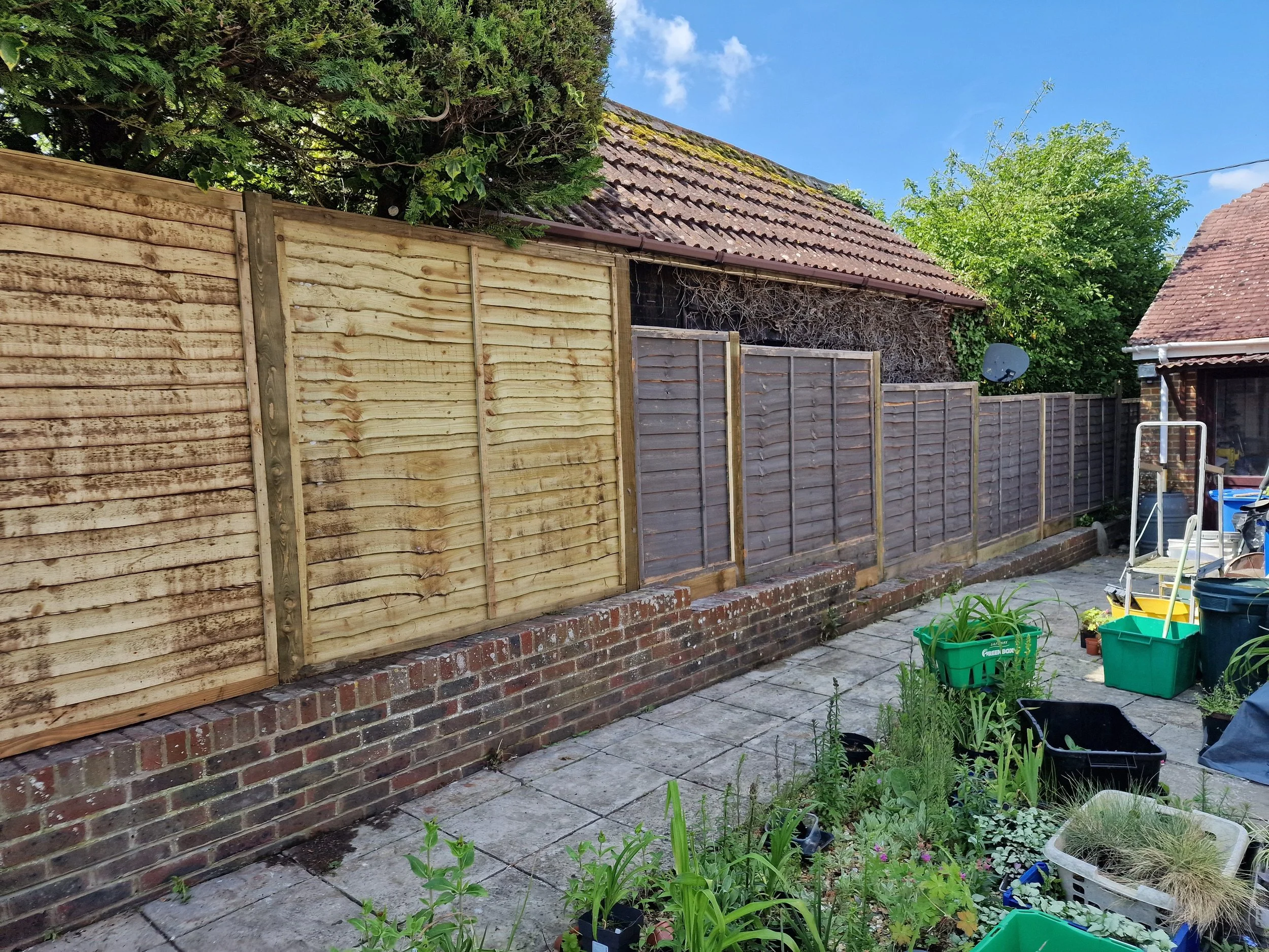 A backyard with a wooden and gray fence, a brick base, and potted plants on a paved patio. There is a house with a tiled sloped roof, a satellite dish, and trees with green foliage against a blue sky.