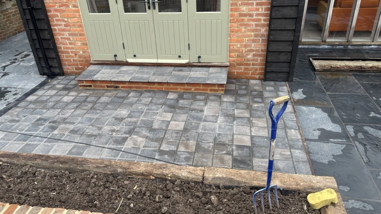 Patio or walkway area with newly installed grey and black stone tiles outside a building with a brick wall and a black wooden wall. A garden fork and a sponge are placed on the ground near the tiles, with a small patch of dirt in the foreground.