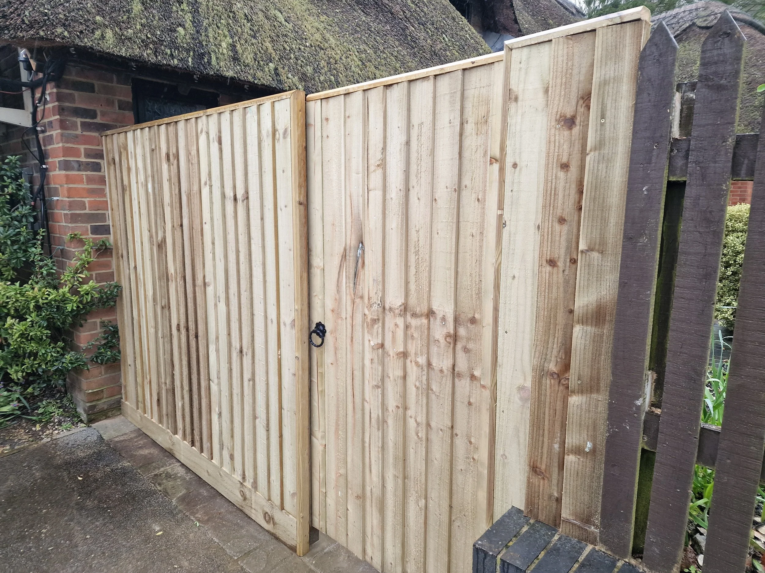 Pre-made wooden fence panels leaning against a partial brick wall with a moss-covered roof, next to some greenery and a wooden picket fence.