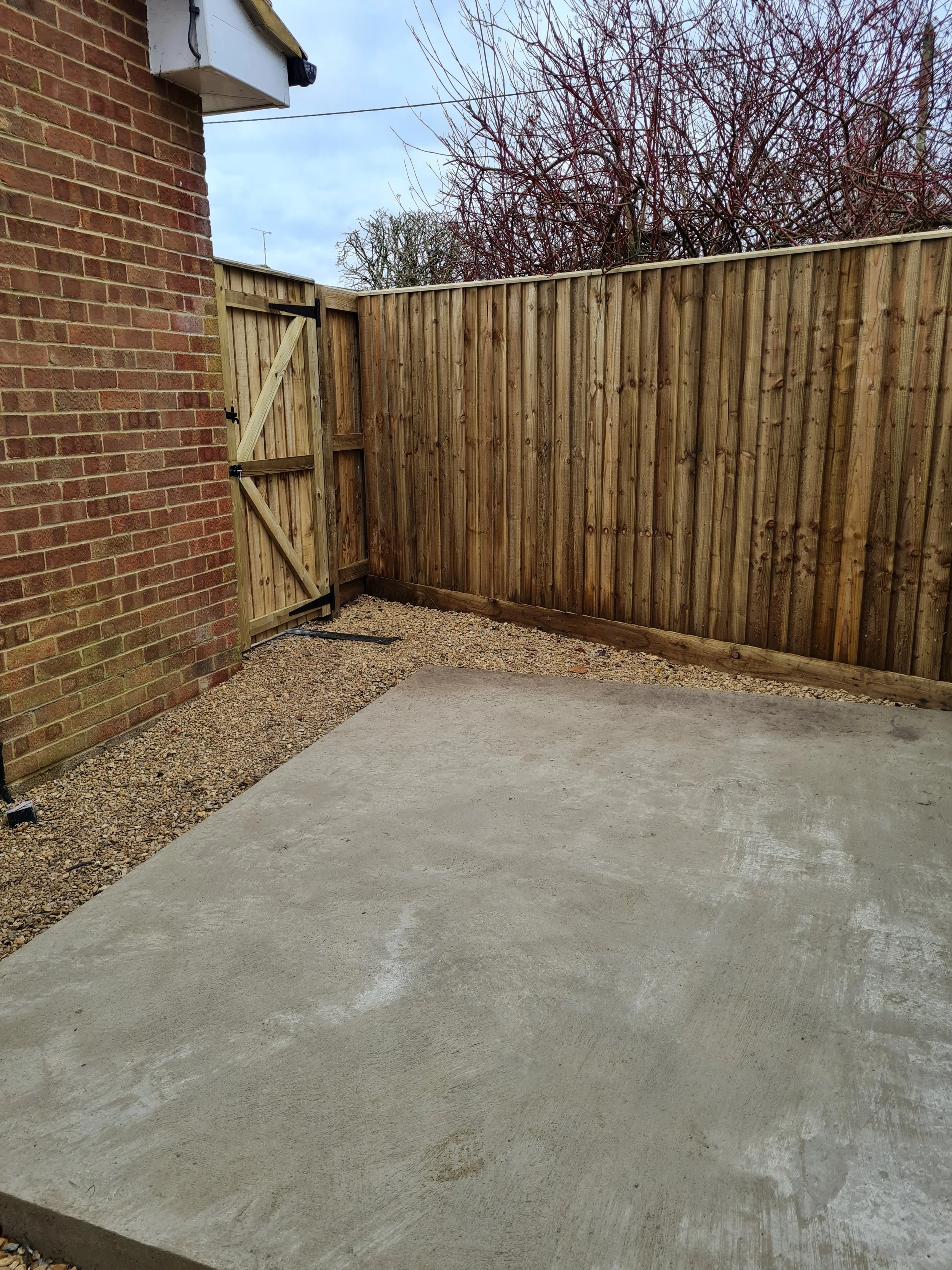 A backyard corner with a red brick house wall, a wooden fence, a small gate, gravel ground, and a concrete patio. The sky is partly cloudy.