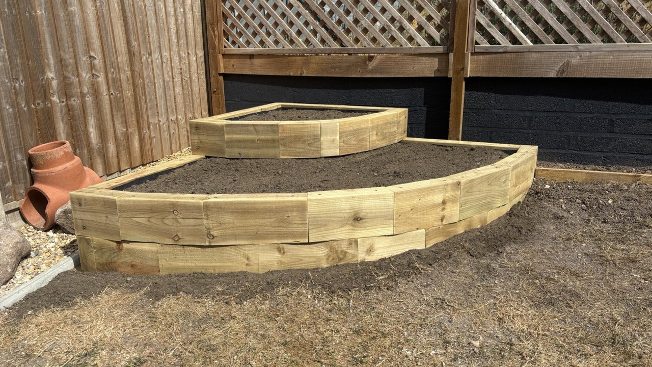 Newly built wooden raised garden beds in a back garden, with dark soil inside, surrounded by a wooden fence and some garden decorations.