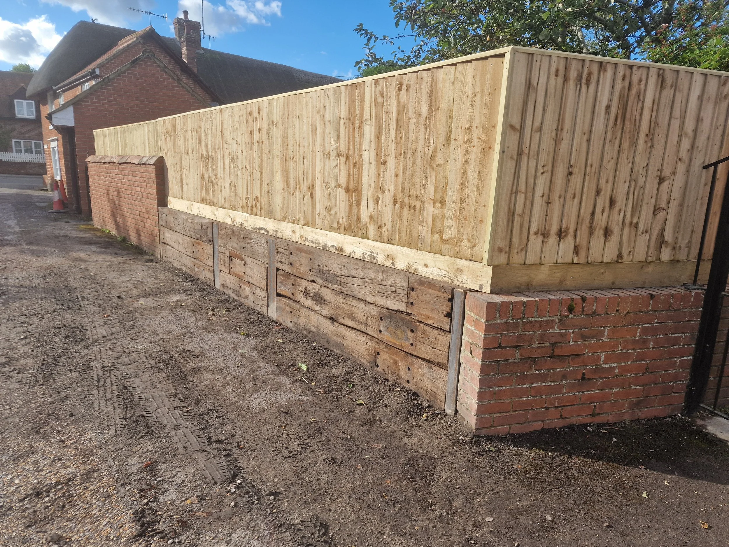 A wooden privacy fence under construction next to a brick wall, with a house and a tree visible in the background on a sunny day.
New railway sleeper retaining wall and Featherboard fencing