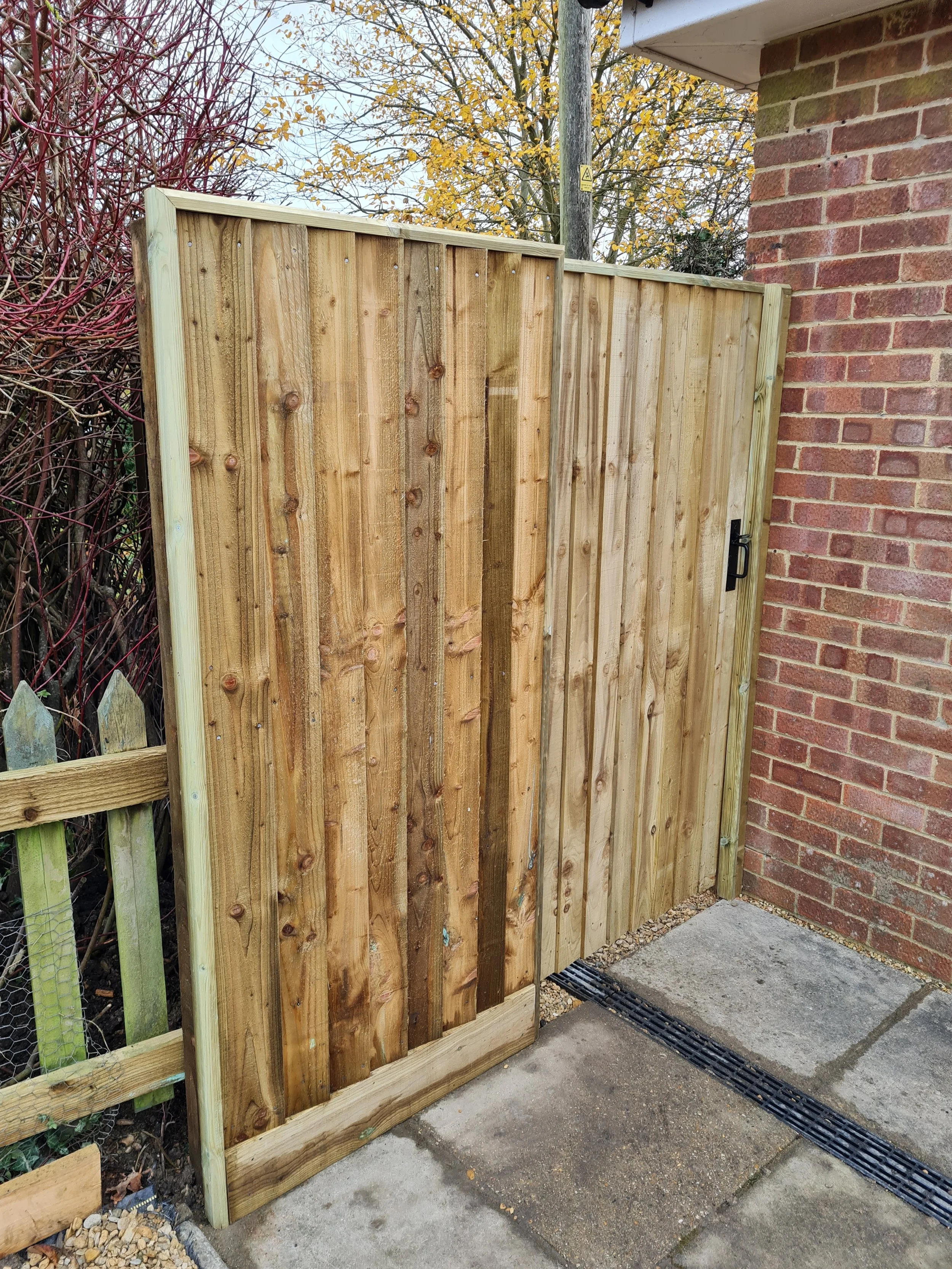 Newly installed wooden fence panel next to a brick wall on a concrete sidewalk with a drainage grate, autumn trees in the background.