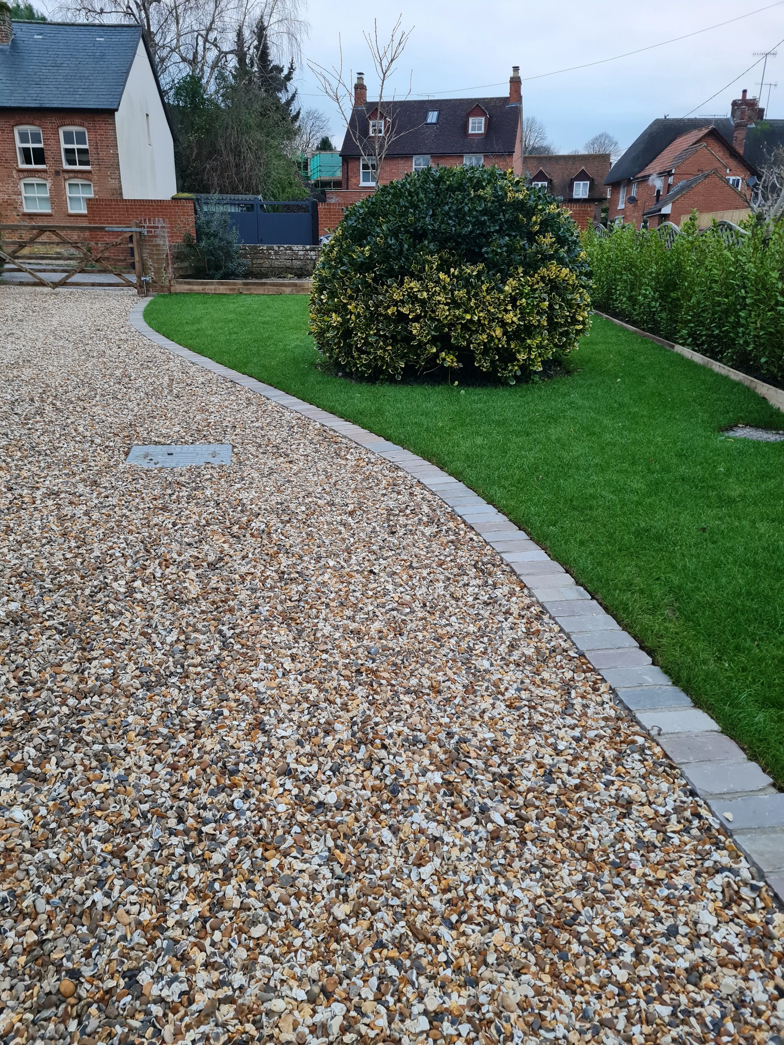 A landscaped yard with a gravel pathway on the left, a neatly trimmed green lawn in the center, and a large rounded bush on the right. In the background, there are traditional brick houses with chimneys and some trees.