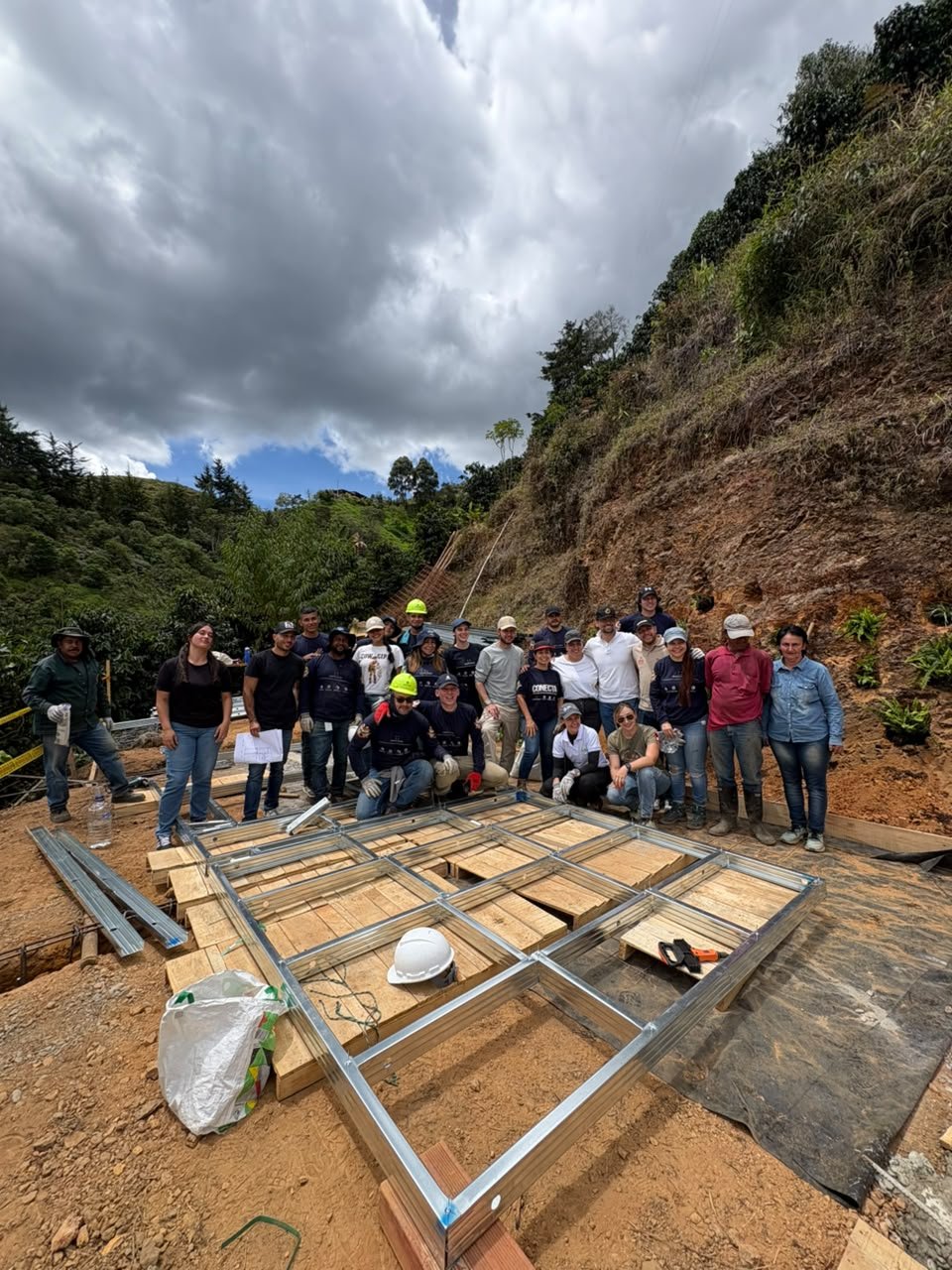 Grupo de personas en un sitio de construcción, algunas con cascos de seguridad, trabajando en la instalación de una estructura de techumbre. Fondo de colinas verdes y cielo nublado.