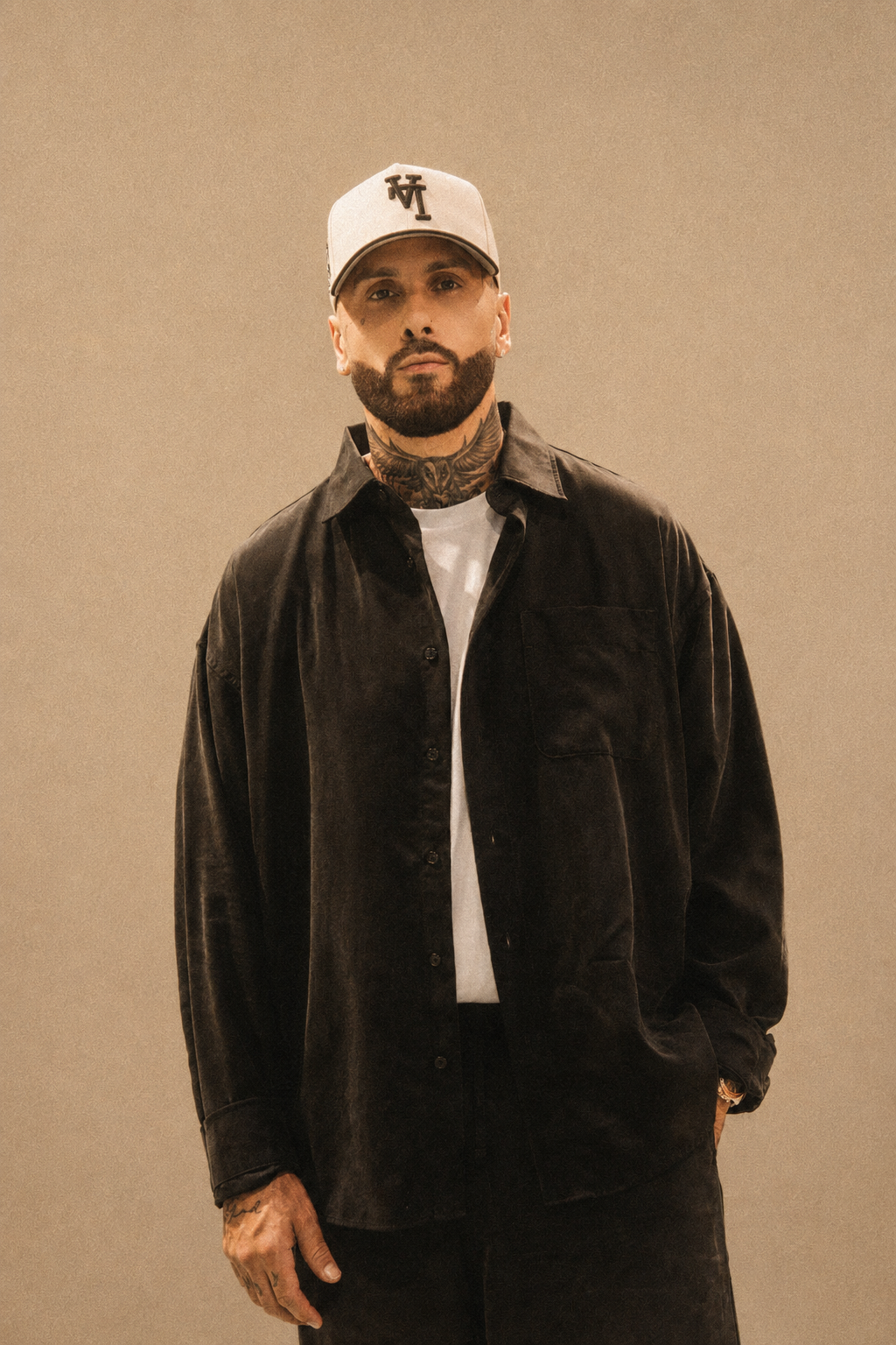 Hombre con barba y tatuajes, lleva gorra blanca con letras negras, camiseta blanca y abrigo negro, posa de frente con fondo beige.