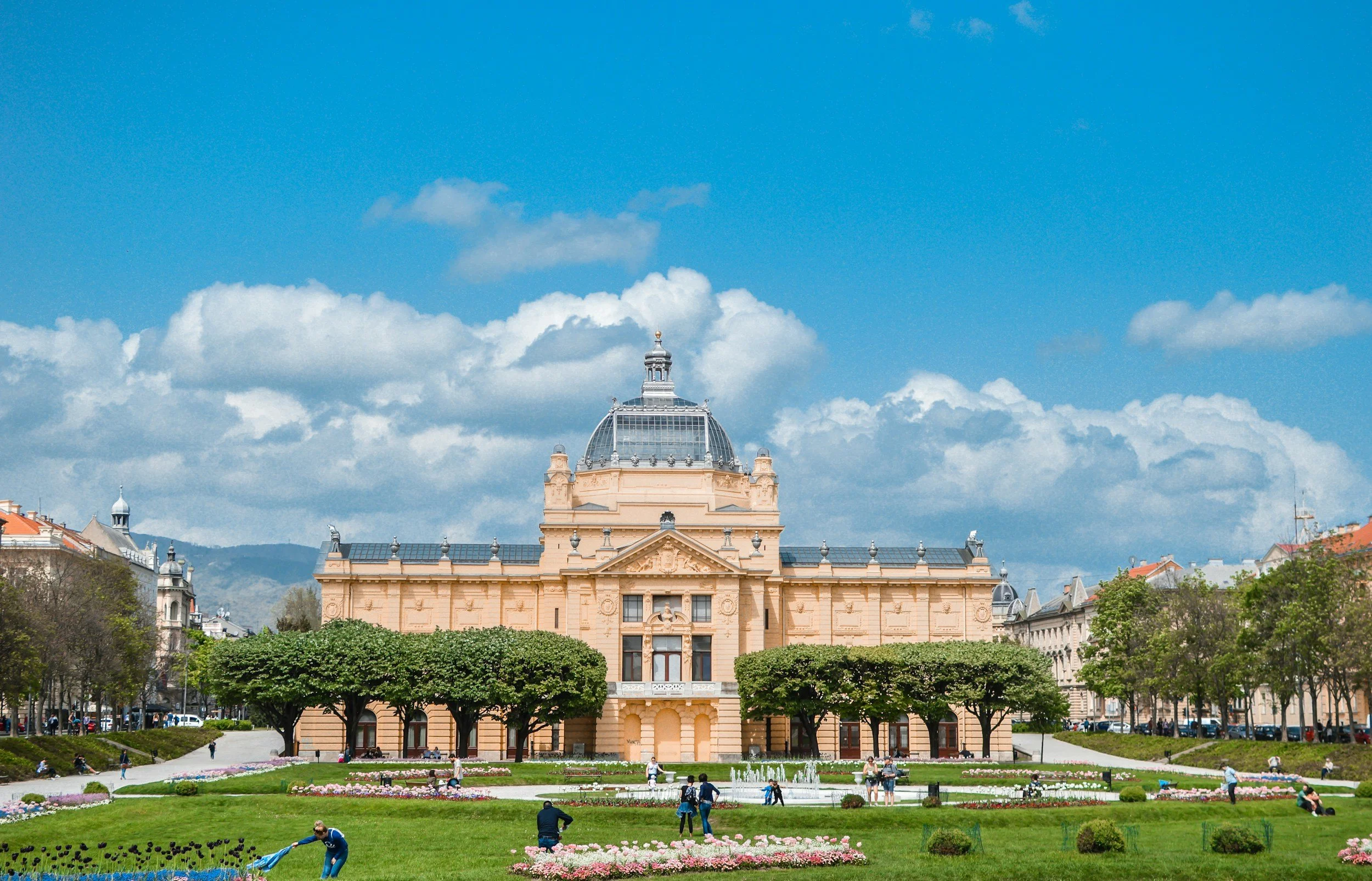 A historic building with a glass dome on top, surrounded by a park with green trees and flowerbeds, blue sky with clouds, and people enjoying outdoors.