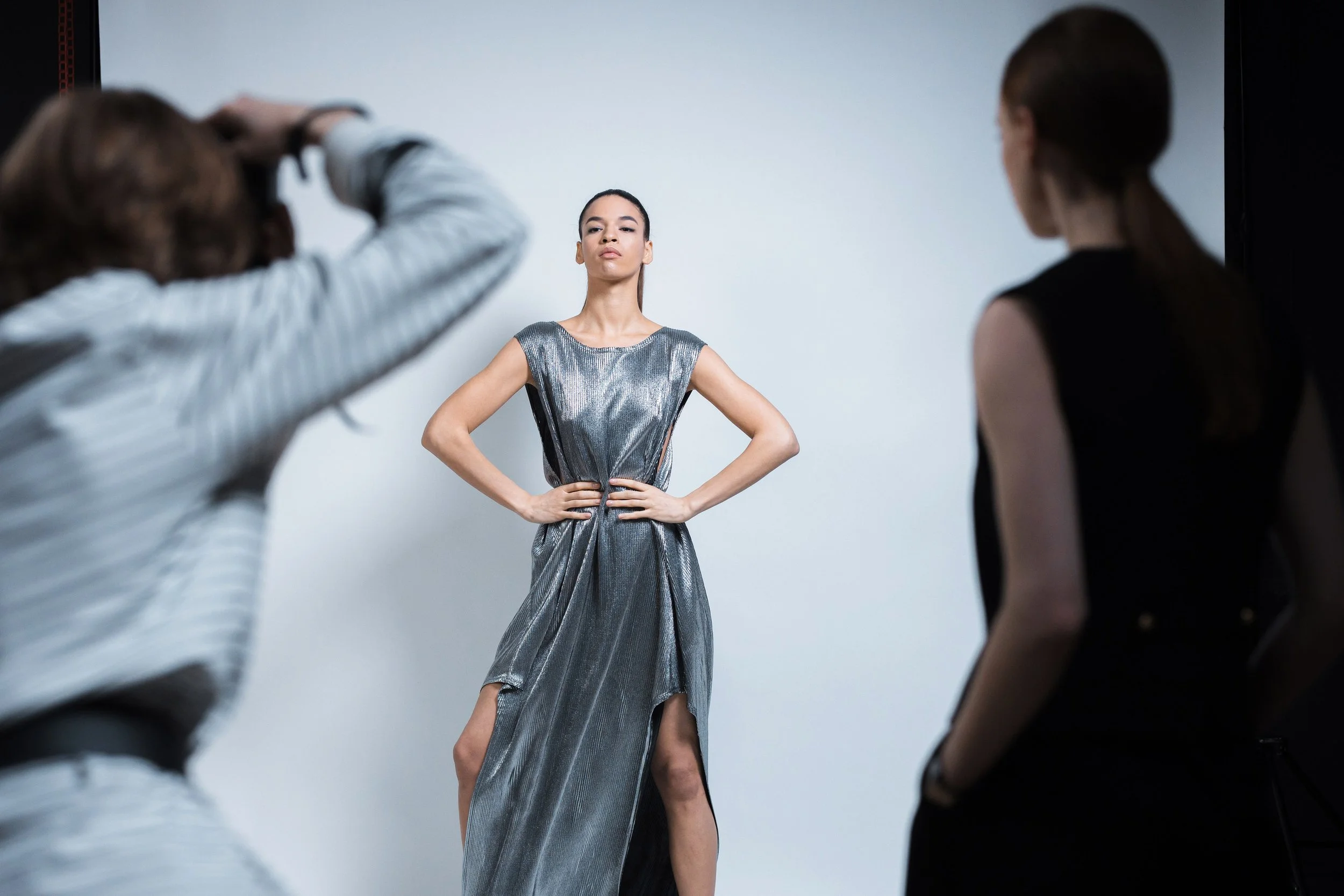 A female model wearing a metallic silver dress with a slit, posing with hands on her hips during a photoshoot with two crew members adjusting the setup.