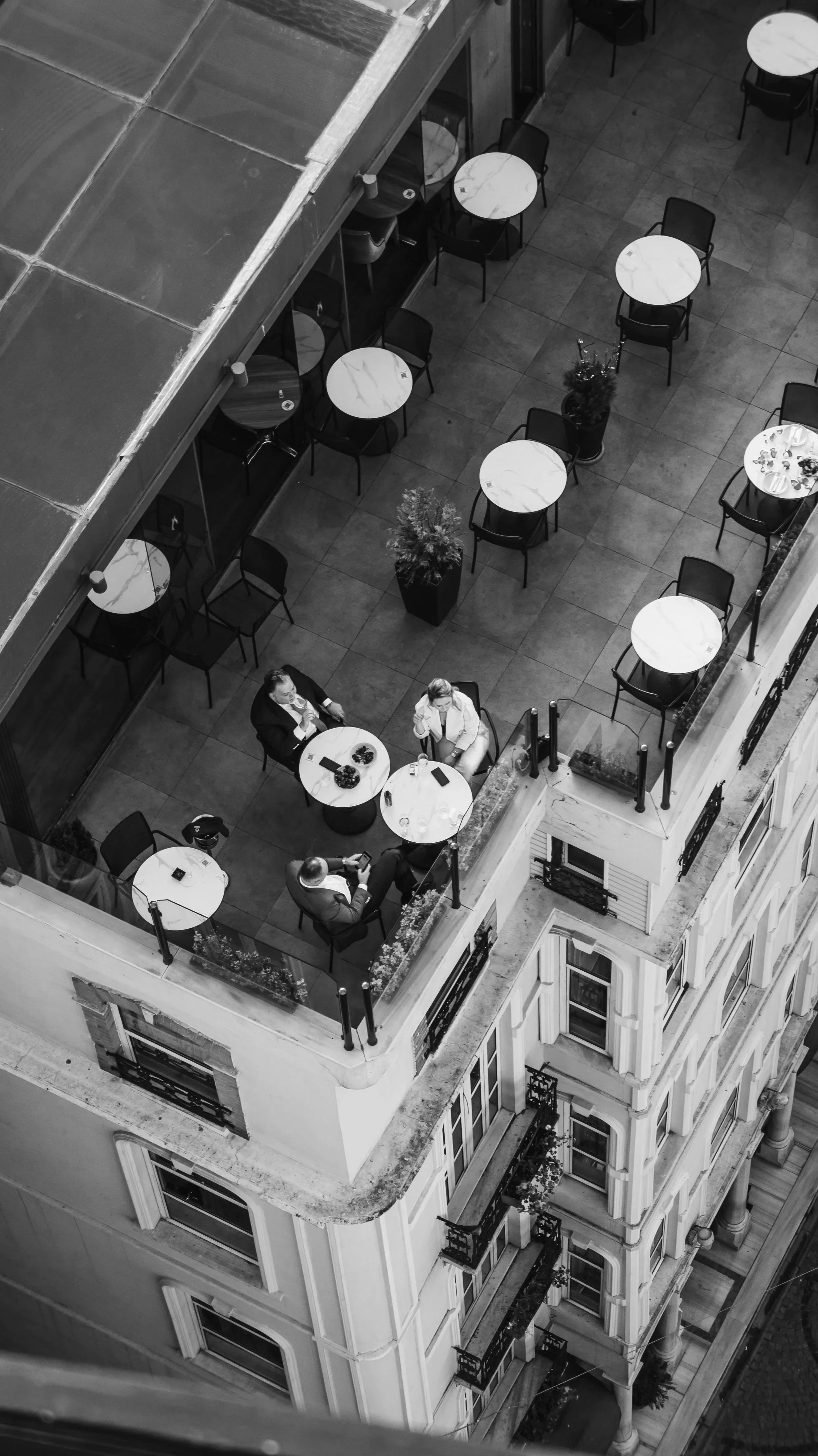 A high-angle black-and-white photo of a rooftop terrace with multiple round tables, some with chairs and a few with people seated and talking, along with potted plants and city buildings in the background.