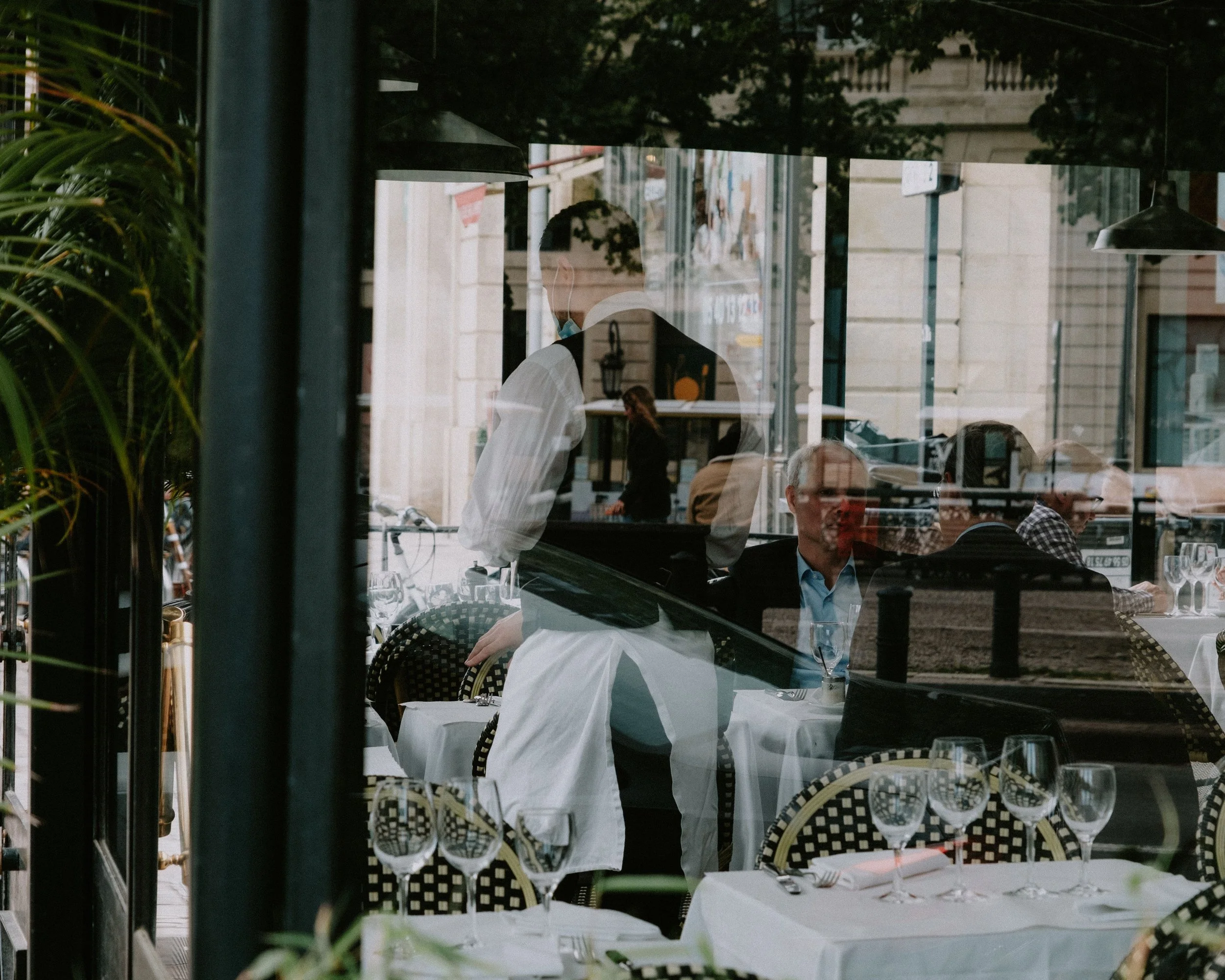 Reflection of people dining at a restaurant through a glass window, with tables set with glasses and checkered chairs outside.