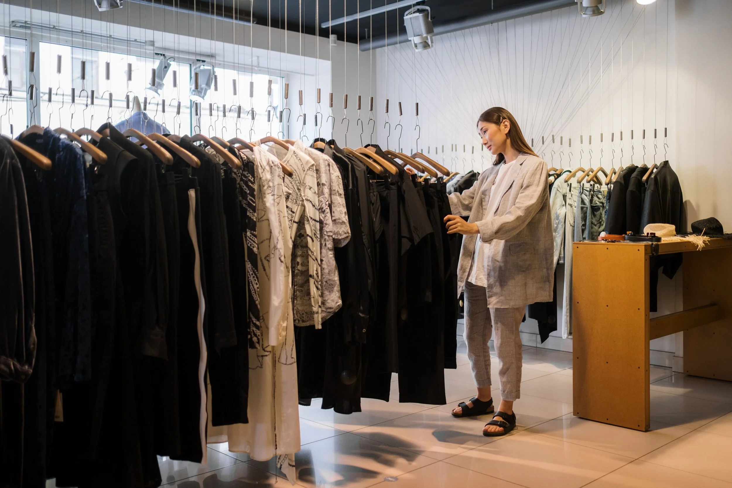 A woman shopping for clothes in a store, browsing through a rack of various tops and pants, with sunlight coming through the windows in the background.
