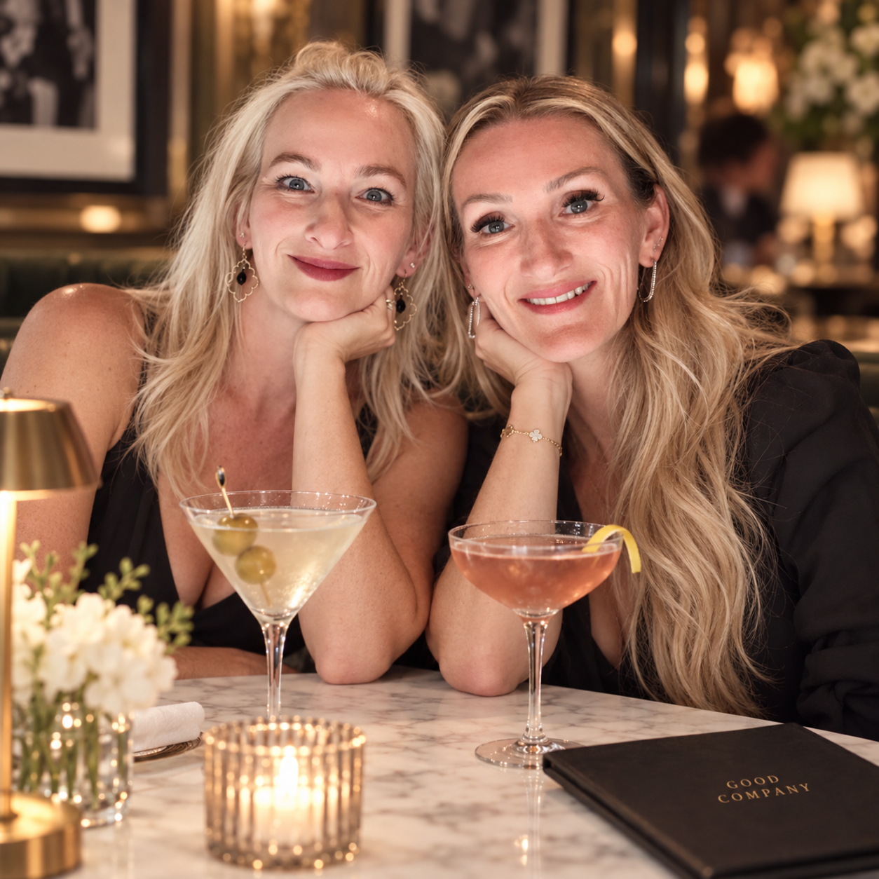 Two women sitting at a table in a restaurant, smiling at the camera. They each have a cocktail in front of them and accessories like earrings and bracelets. The table has a small floral arrangement and a candle. The background is warmly lit.