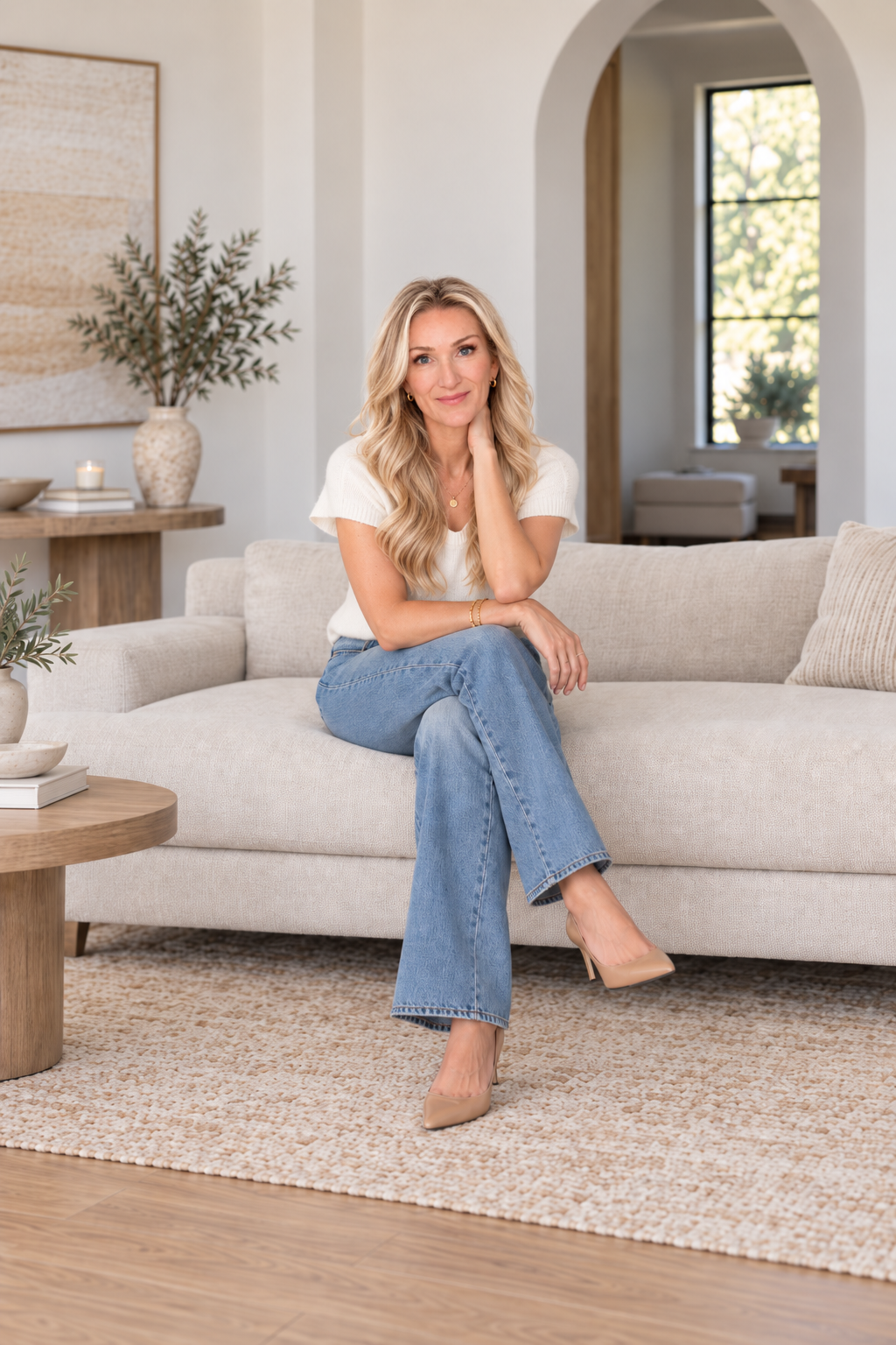 A woman sits on a light-colored sofa in a modern, cozy living room with natural light, plants, and minimal decor.