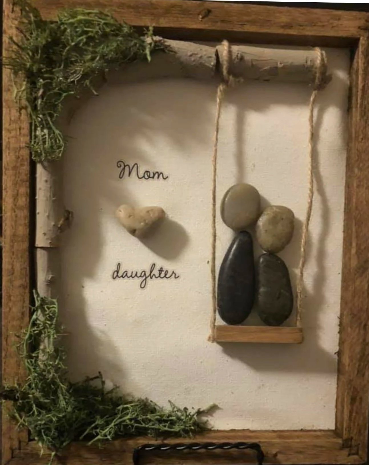 A framed decorative display with a white background. On the left side, the words "Mom" and "daughter" are written with small rocks arranged as hearts. On the right side, the words are written in lines with two stones and a spoon representing the figures of a mother and daughter holding hands, with the spoon's handle forming the arm.