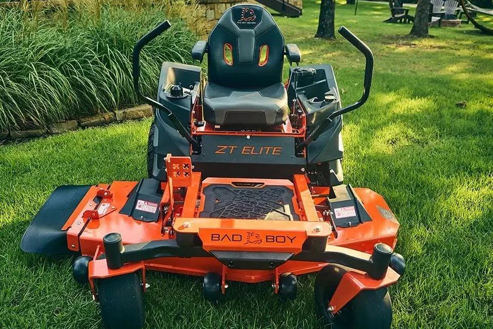 A zero-turn riding lawn mower on a grassy yard with trees and outdoor seating in the background.
