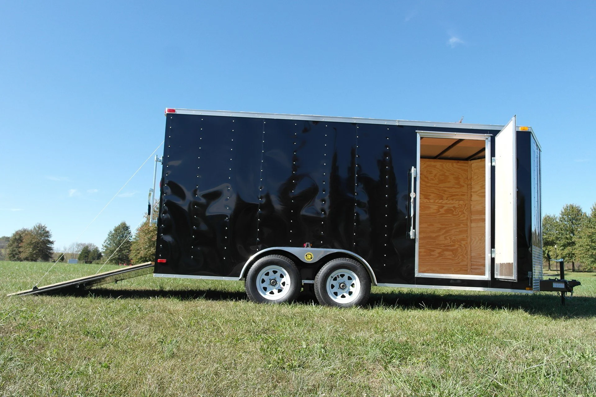 Black enclosed cargo trailer with open side door, parked on grassy field with trees in the background, under a clear blue sky.