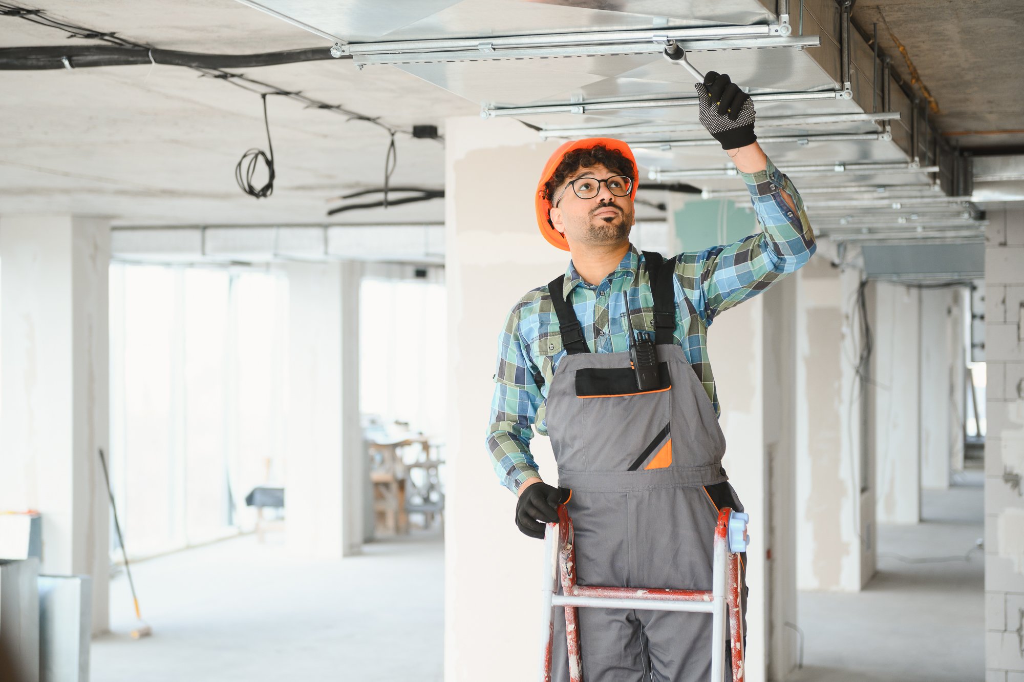 A man wearing a plaid shirt, safety vest, glasses, and orange safety helmet is standing on a ladder while working on ceiling tiles in a construction site. He is using a tool to adjust the ceiling structure.