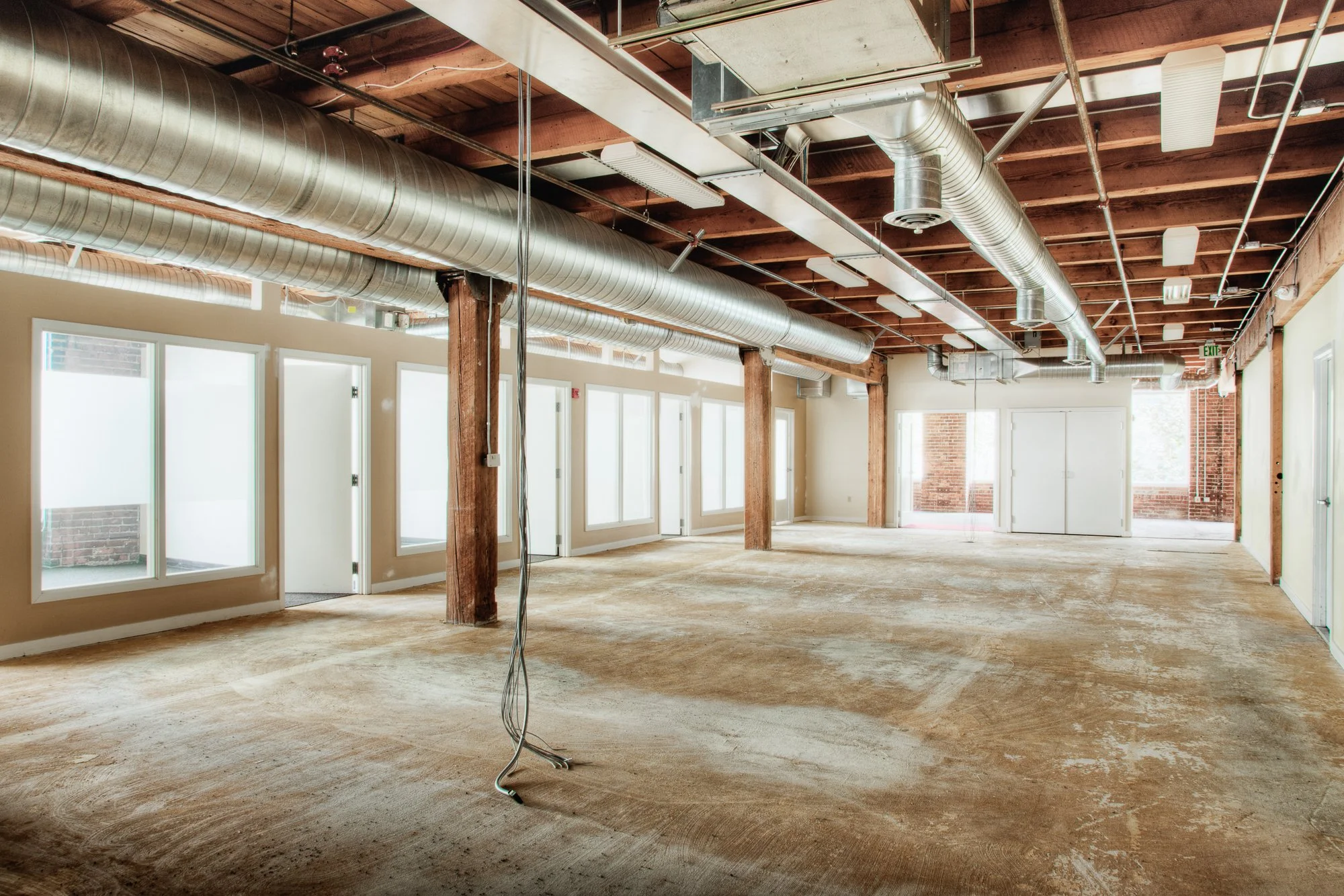Empty commercial or office space under construction with exposed ductwork, electrical wiring, wooden ceiling beams, and large windows.