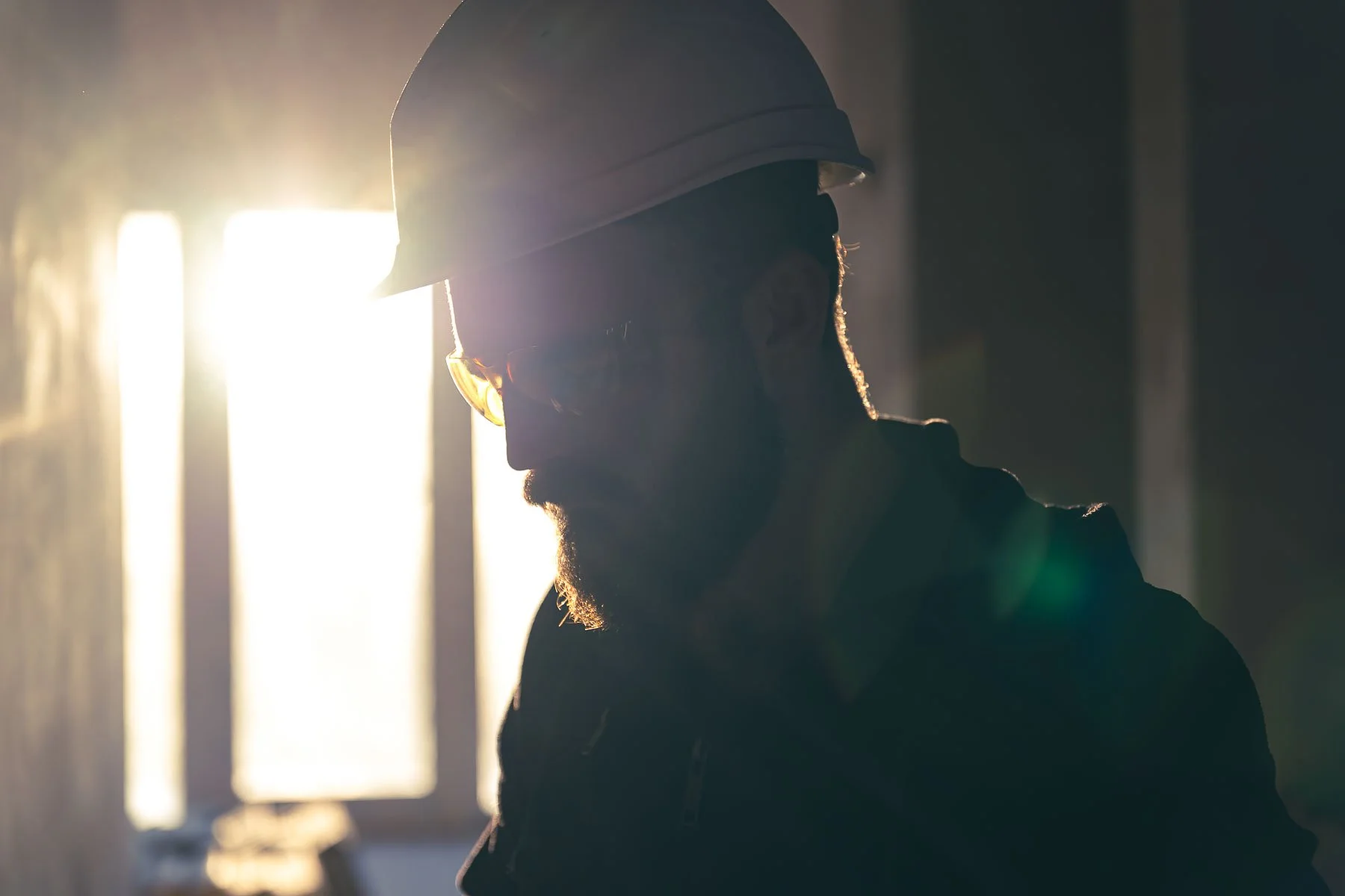Silhouette of a bearded man wearing safety glasses and a hard hat, backlit by bright sunlight coming through windows.