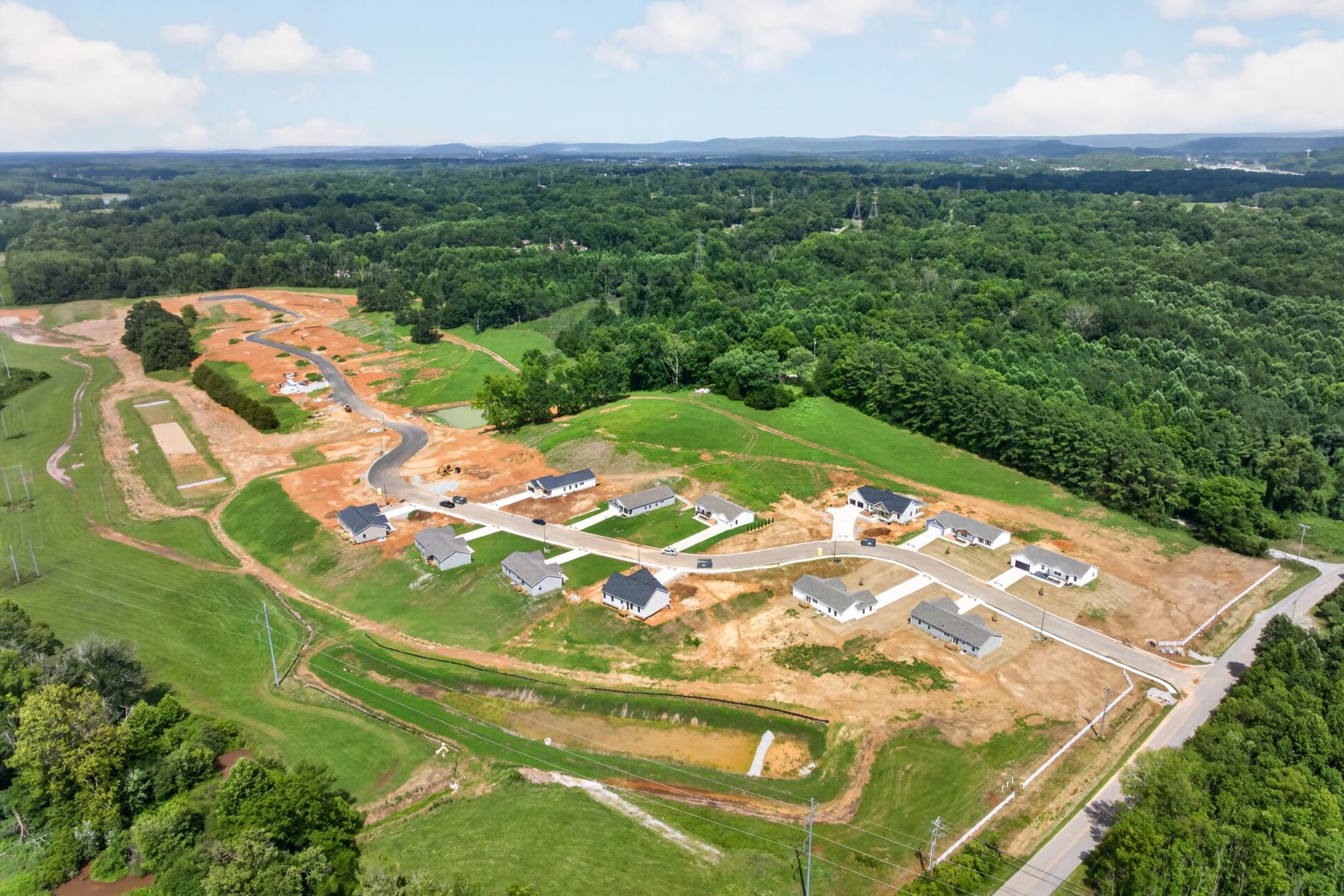 Aerial view of a residential development under construction with cleared soil and infrastructure, surrounded by dense green forests and hills in the distance.