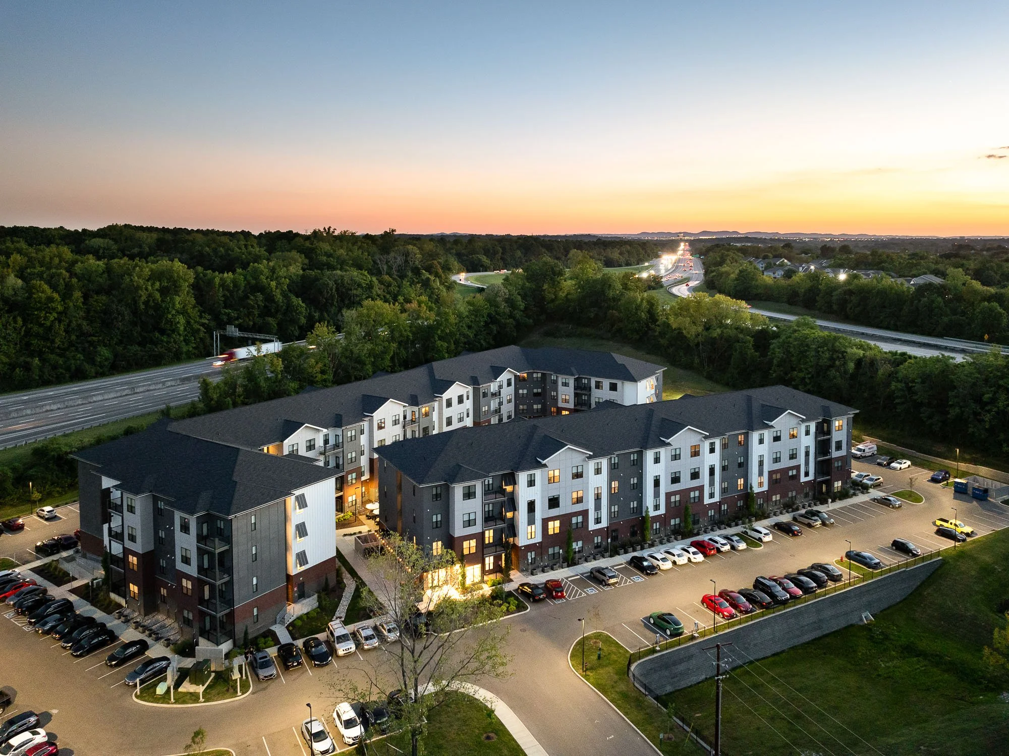 Aerial view of residential apartment complex with parking lot, trees, and surrounding highway at dusk, with a colorful sunset in the sky.