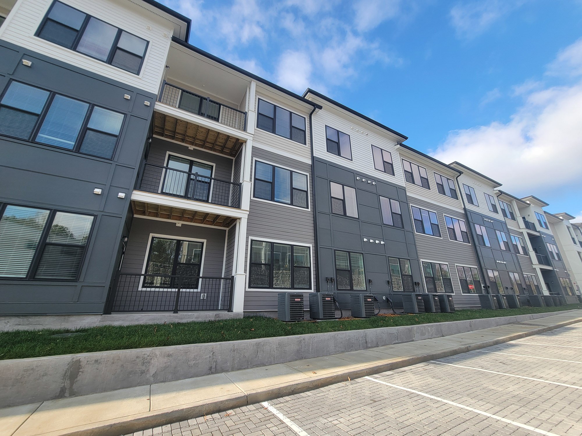 Exterior view of a modern multi-story apartment building with multiple windows, balconies, and air conditioning units on the ground level, under a partly cloudy sky.