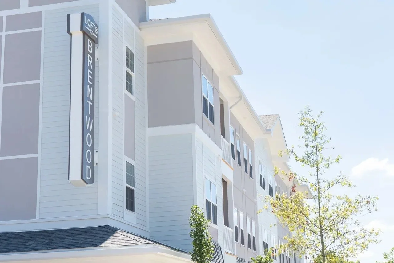 A multi-story apartment building with a sign that reads "LOFTS AT BRUNSWOOD," featuring white and gray exterior walls, multiple windows, and some trees nearby under a clear blue sky.